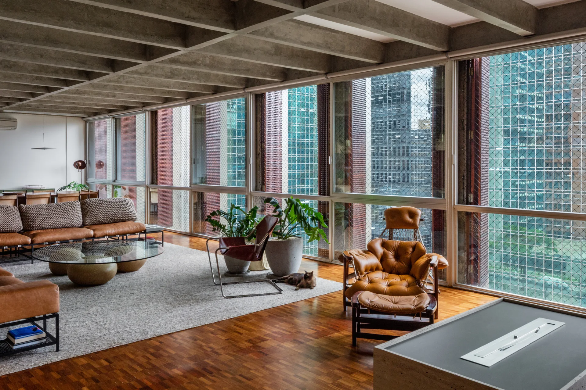 A mid-century modern living room features an exposed concrete grid ceiling, warm leather furniture, a glass coffee table, and large windows.