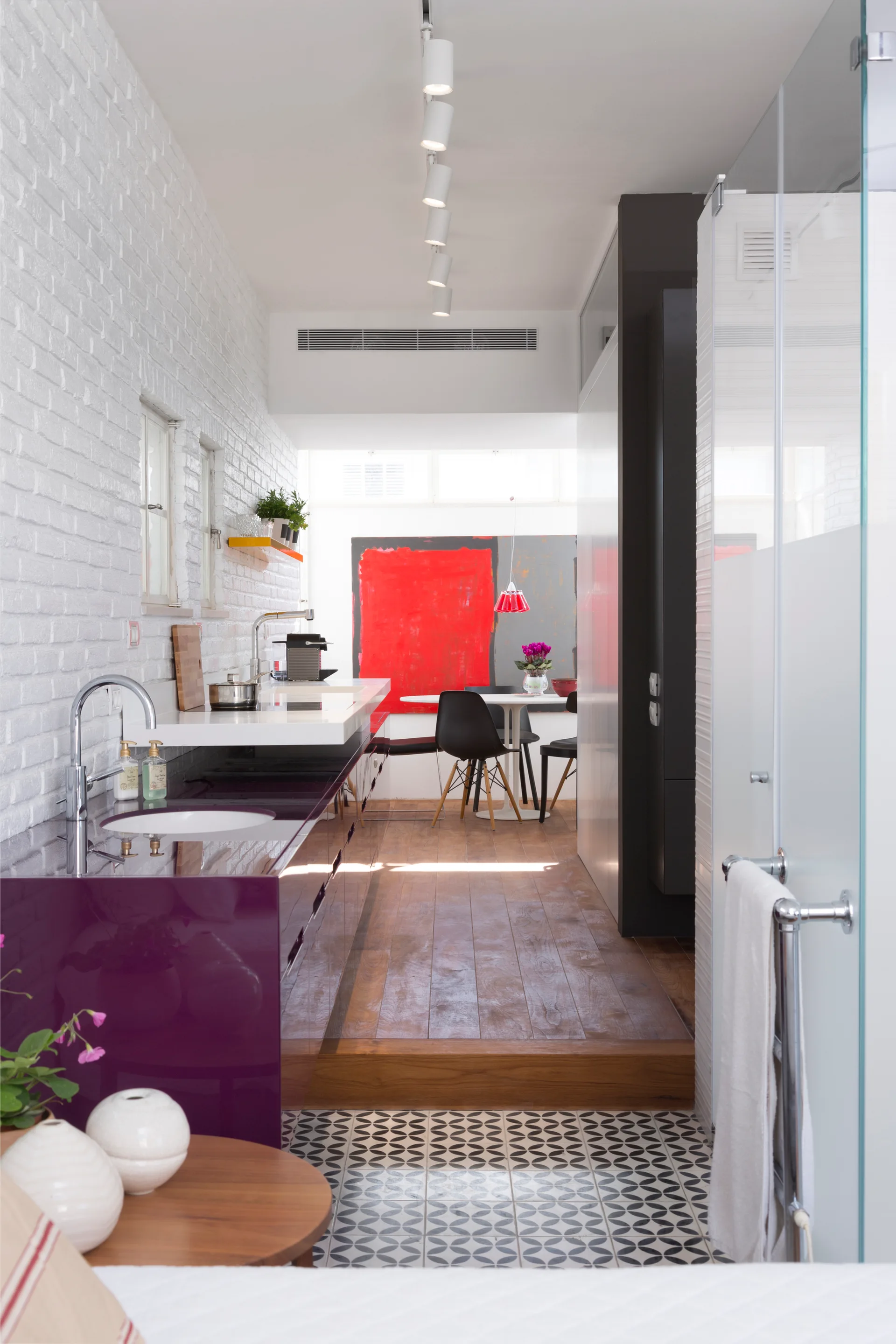 A narrow modern kitchen features glossy purple cabinets against a white brick wall, transitioning to a raised dining area with a bold red painting.