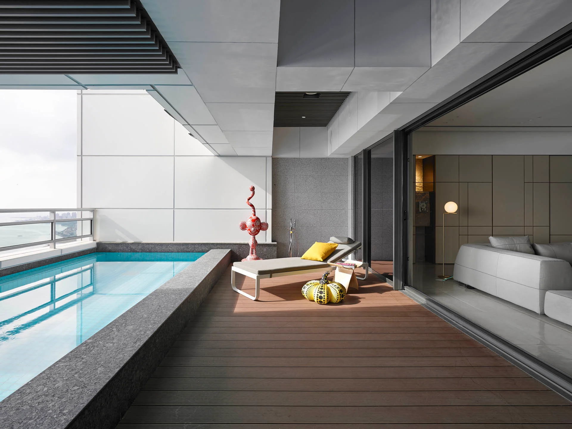 A modern outdoor terrace features a narrow swimming pool, a single chaise lounge with a yellow pillow, and wood plank decking next to large glass doors.