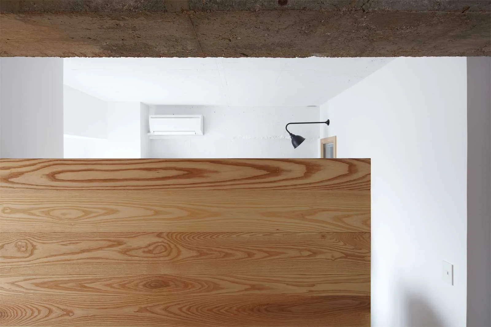 A minimalist white room features a light wood grain partition wall beneath an exposed concrete ceiling beam with a black wall sconce.