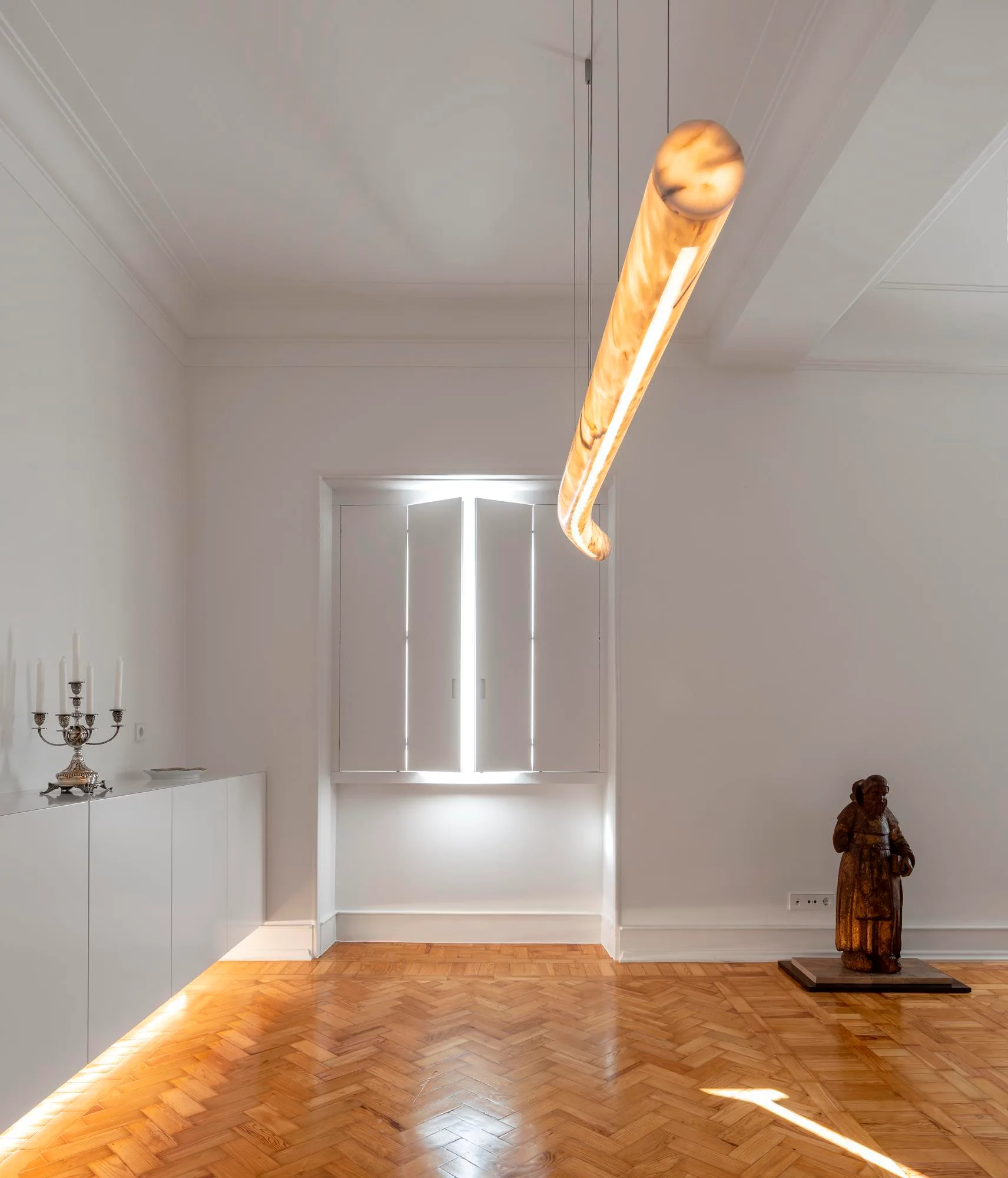 A minimalist room featuring a warm wood herringbone floor, a low white cabinet with base lighting, and a suspended tubular light fixture.