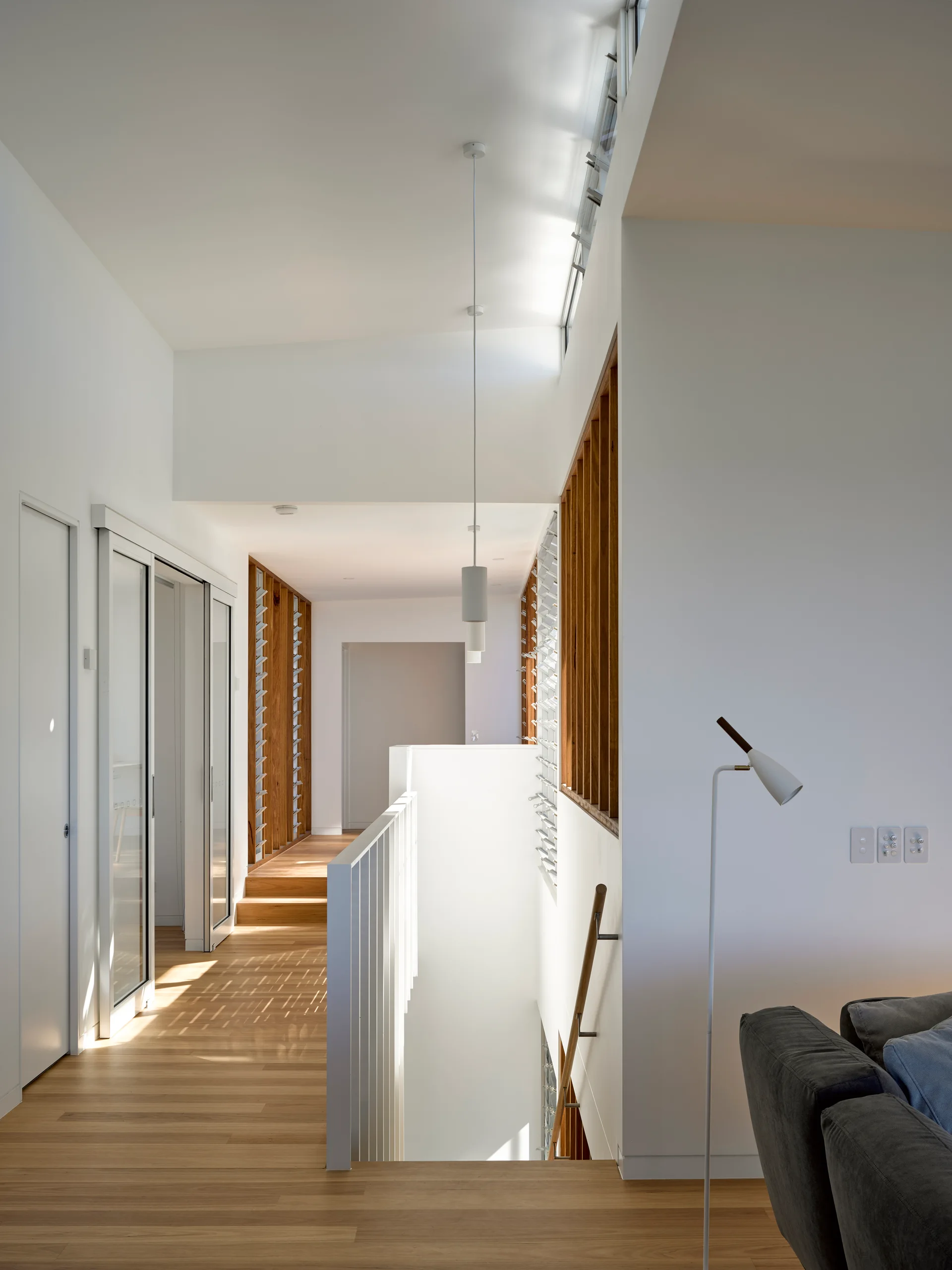 A bright modern hallway and stairwell landing featuring light wood floors, a white metal railing, and a wall of vertical timber slats below high windows.