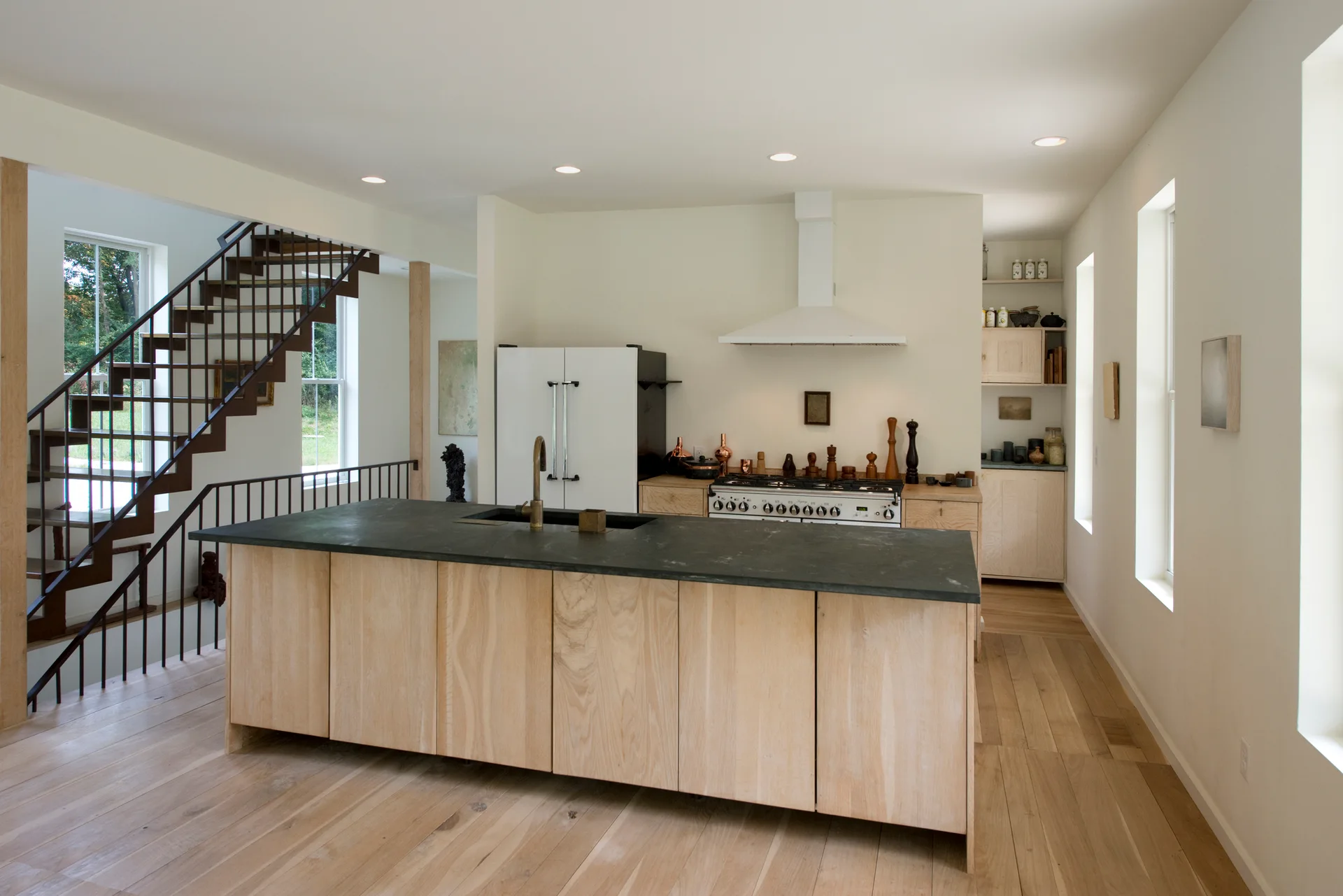 An open-concept kitchen features a light wood island with a dark stone countertop next to a black metal staircase and a white refrigerator.