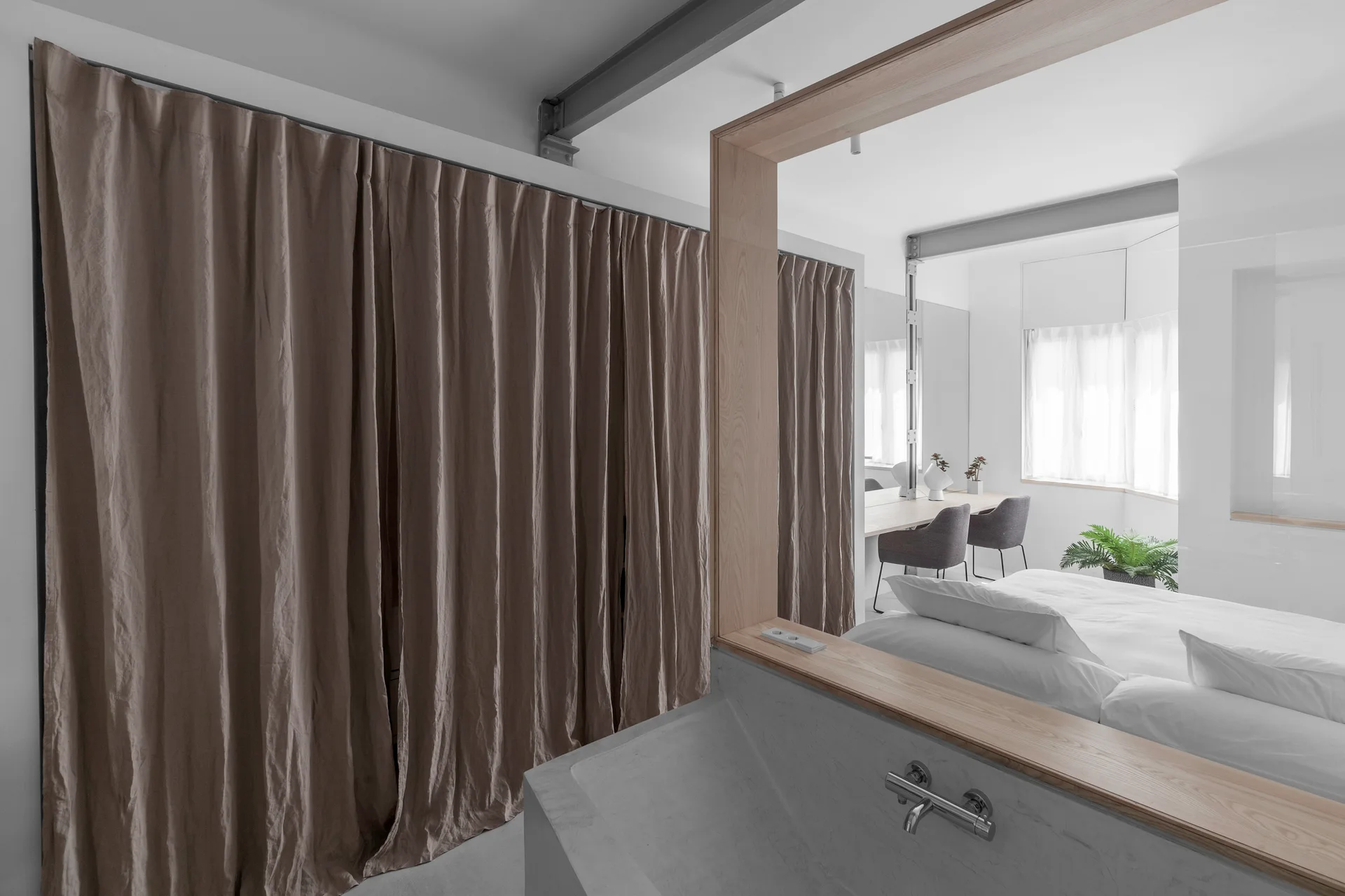 A modern bedroom featuring a bed with white linens, grey curtains, a long wood desk, and a light wood-framed partition in the foreground.
