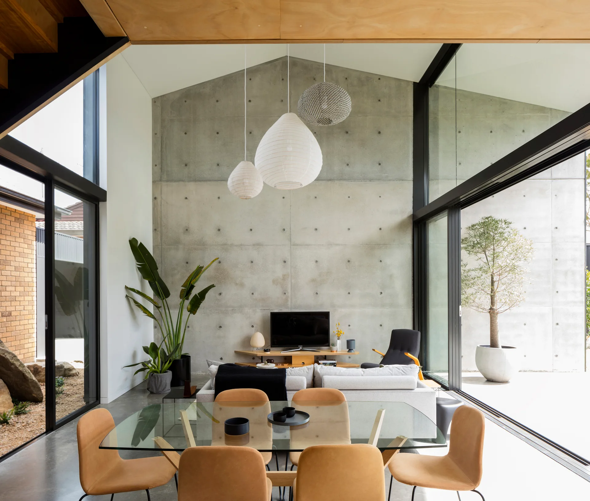 A modern open-concept dining and living space features a glass-topped dining table with tan chairs, a grey sofa, and a large concrete feature wall.
