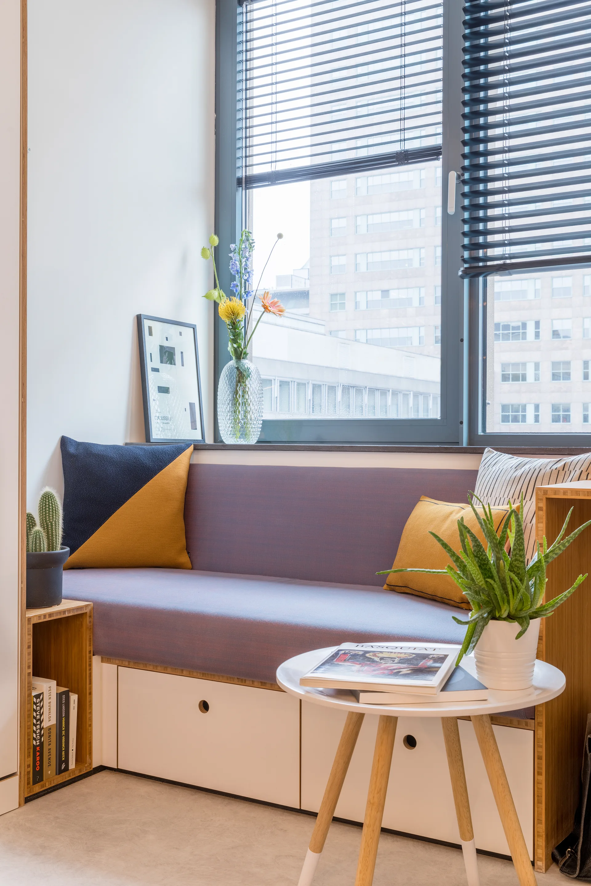 A light purple built-in window seat features lower white storage drawers, colorful accent pillows, and a round wooden side table with potted plants.