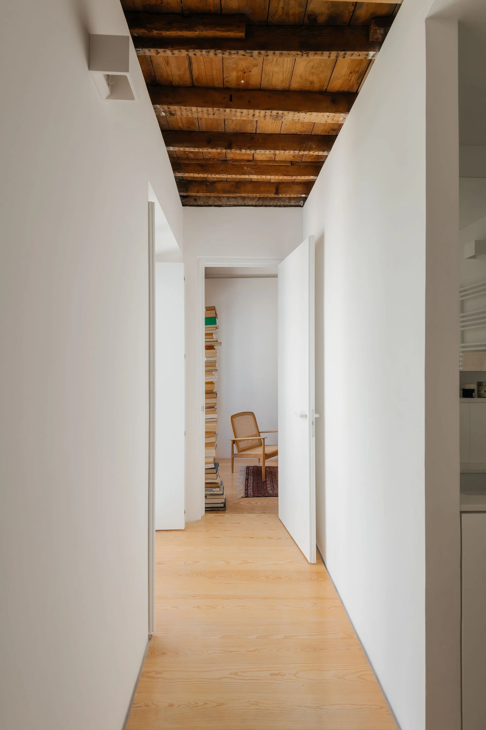 A narrow white hallway with light wood floors and an exposed beam ceiling leads to an open doorway showing a rattan chair and a stack of books.