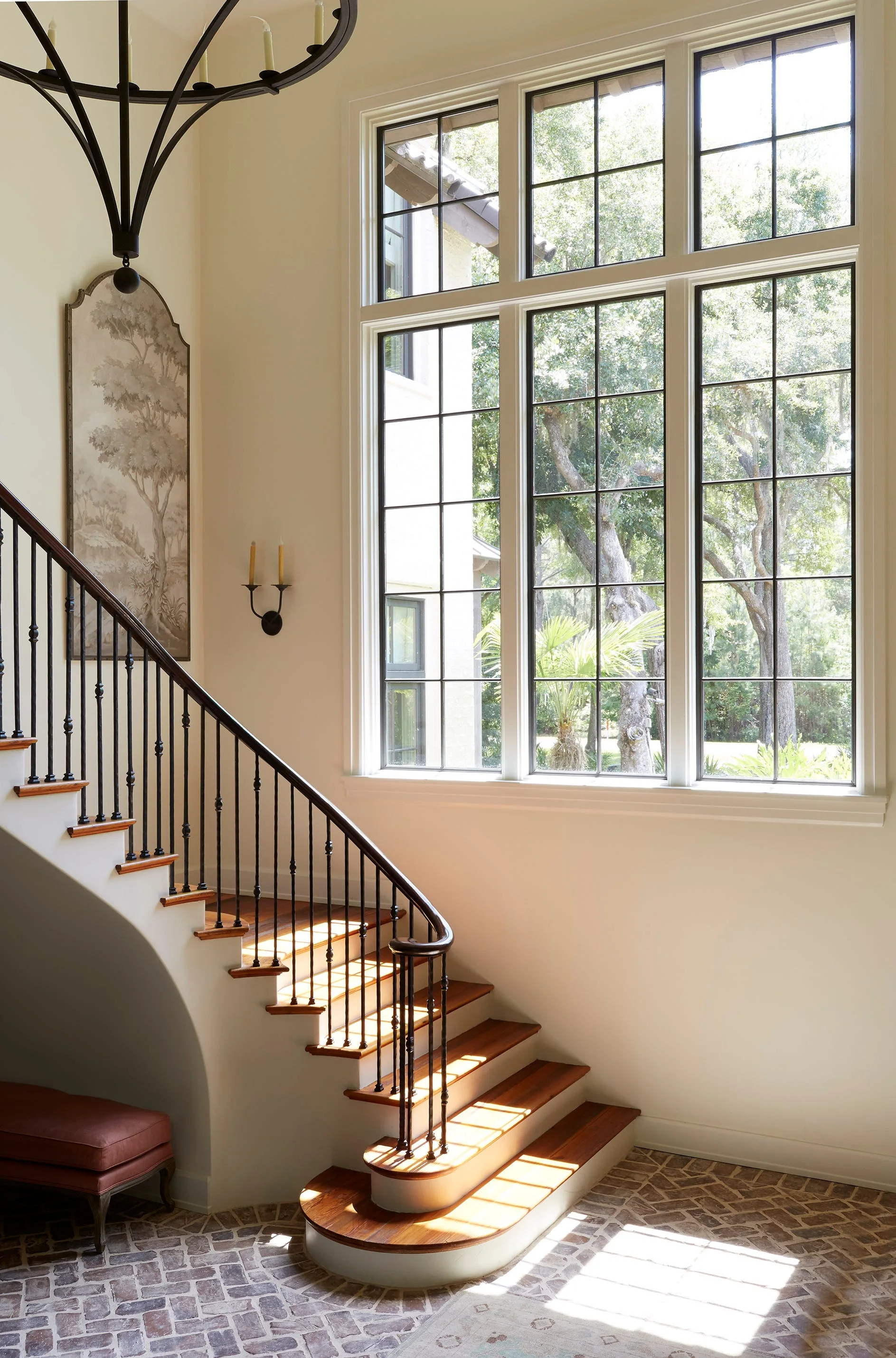 A sunlit foyer features a curved wooden staircase with an iron railing, a herringbone brick floor, and a large multi-pane grid window.