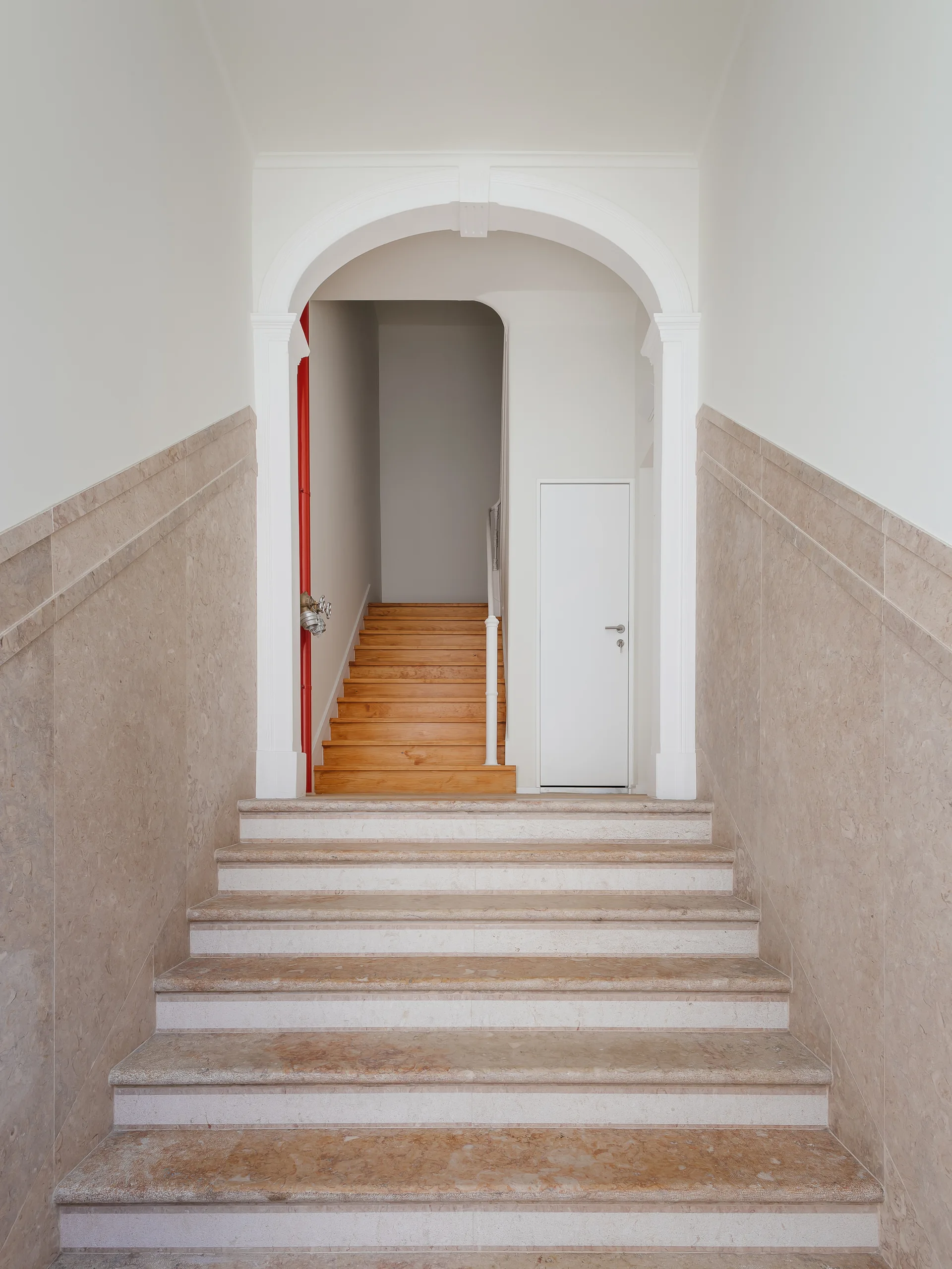 A stone staircase with beige marble treads leads up to a classical white archway, revealing a second flight of light wood steps.