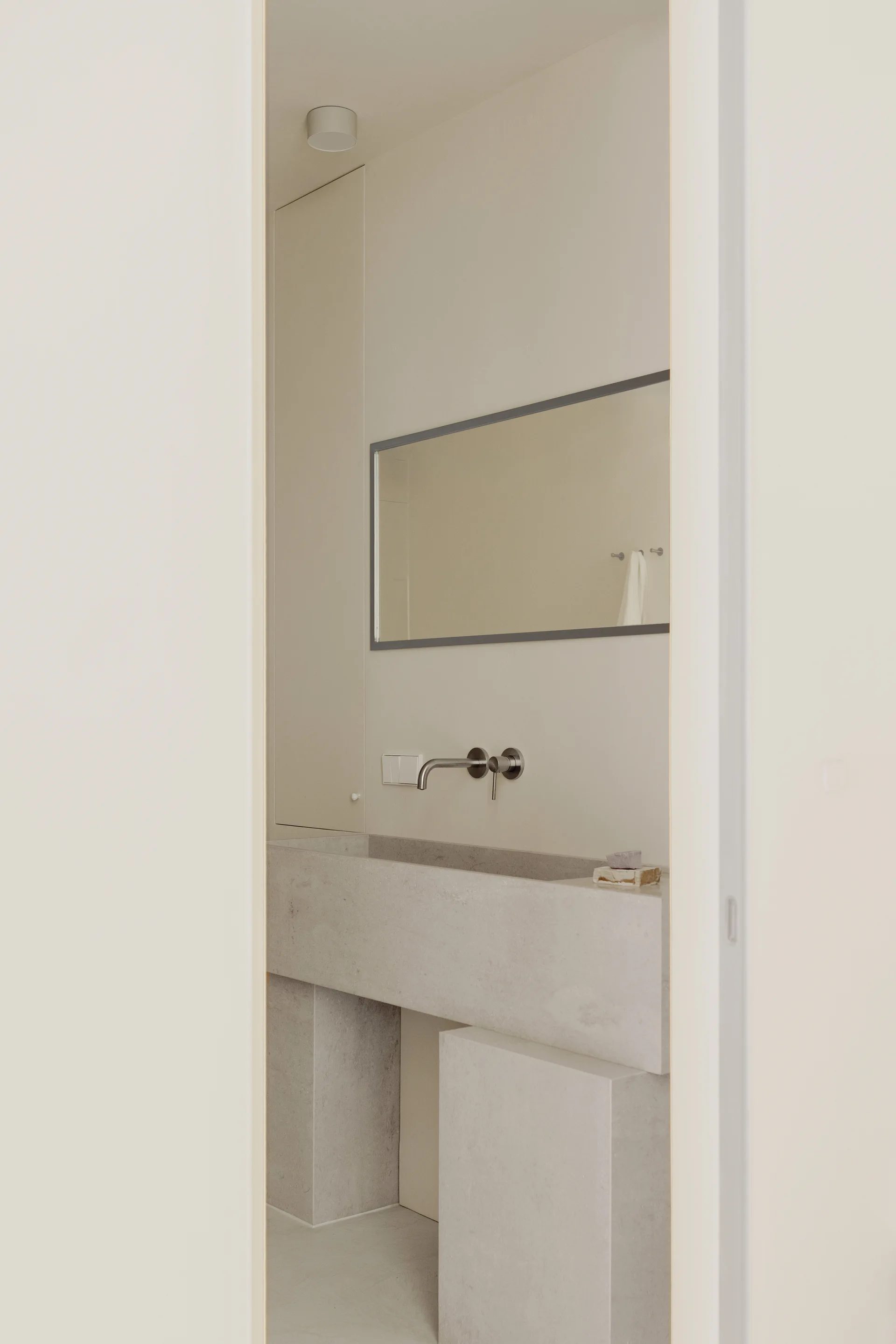 A minimalist powder room featuring a large grey monolithic stone basin, a wall-mounted metal faucet, and a rectangular mirror against beige walls.