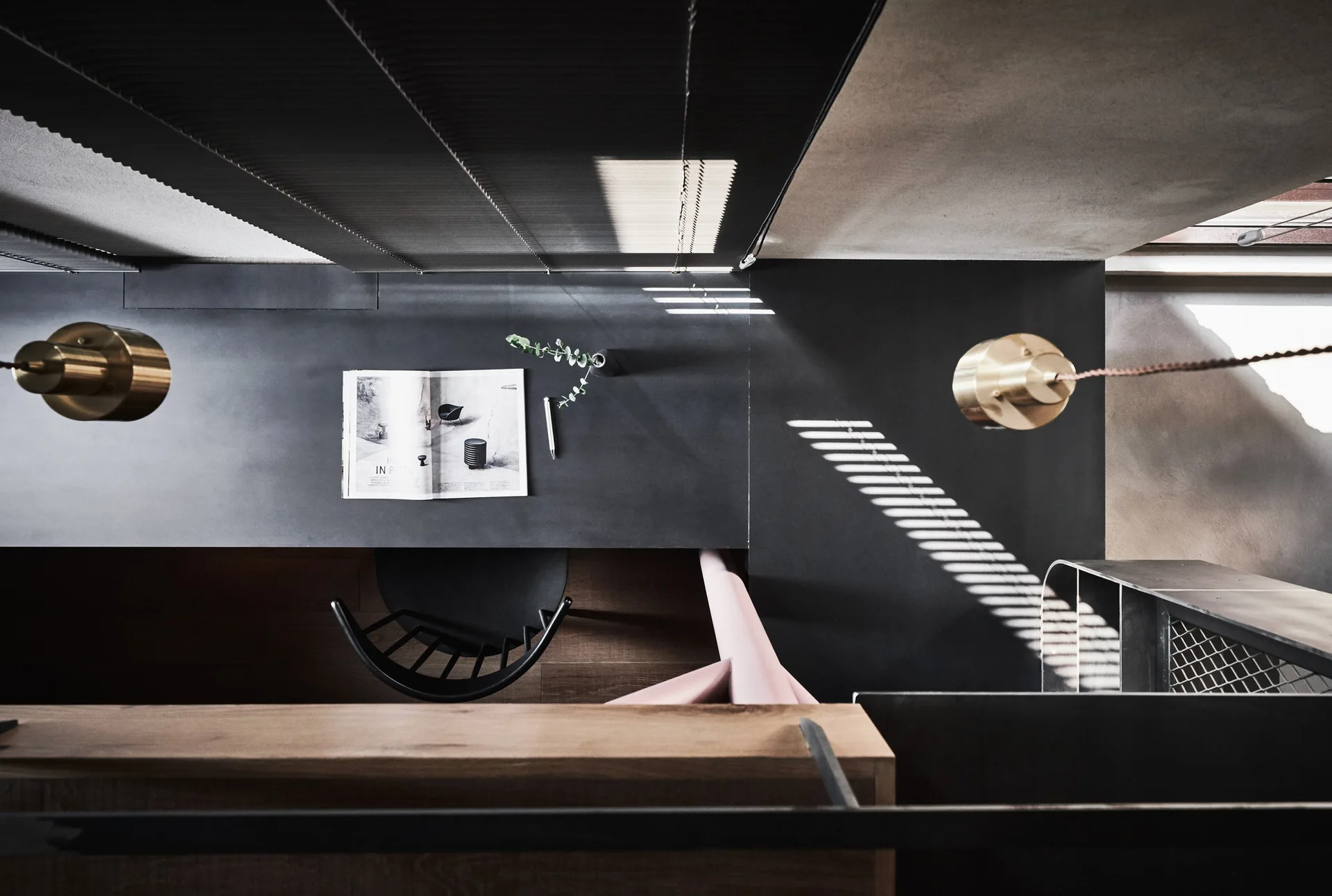 An overhead view of a minimalist black desk with an open book, a black chair, and hanging brass cylinder pendant lights.