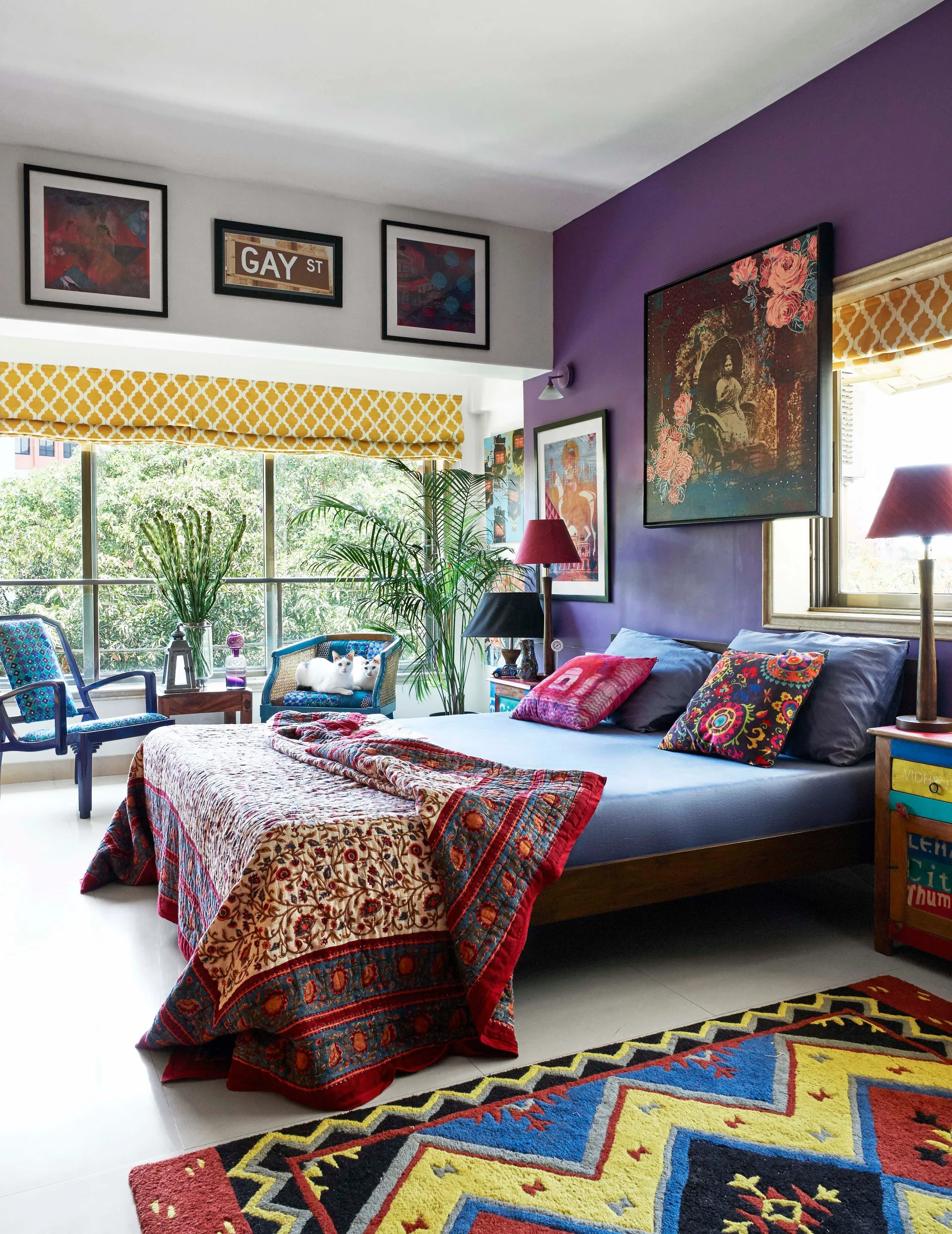 An eclectic bedroom features a purple accent wall behind a wooden bed, patterned quilts, and yellow geometric window valances near a large window.