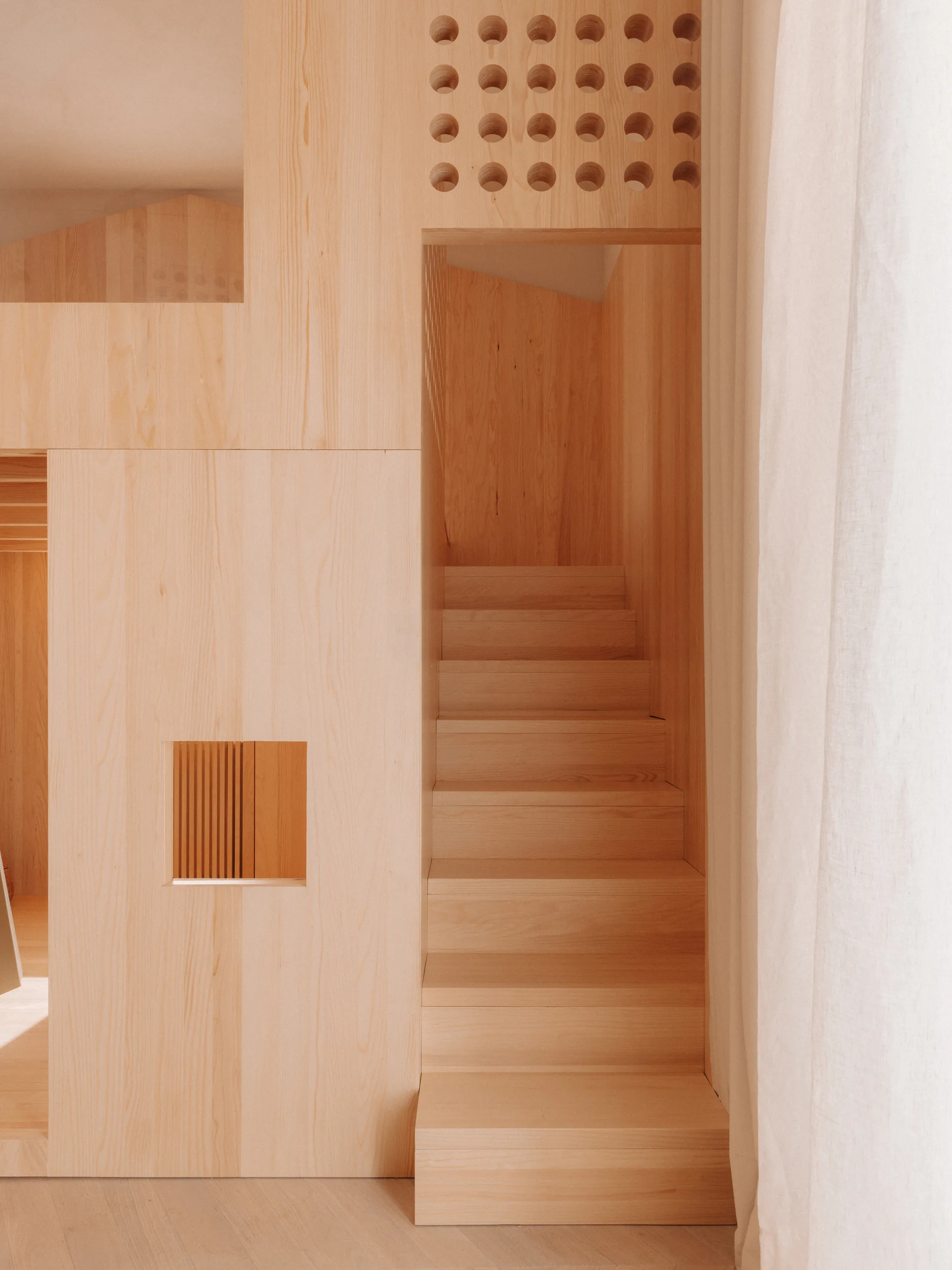 A built-in staircase made of light-toned wood features a matching wood wall paneled structure with a grid of circular cutouts and a small square opening.
