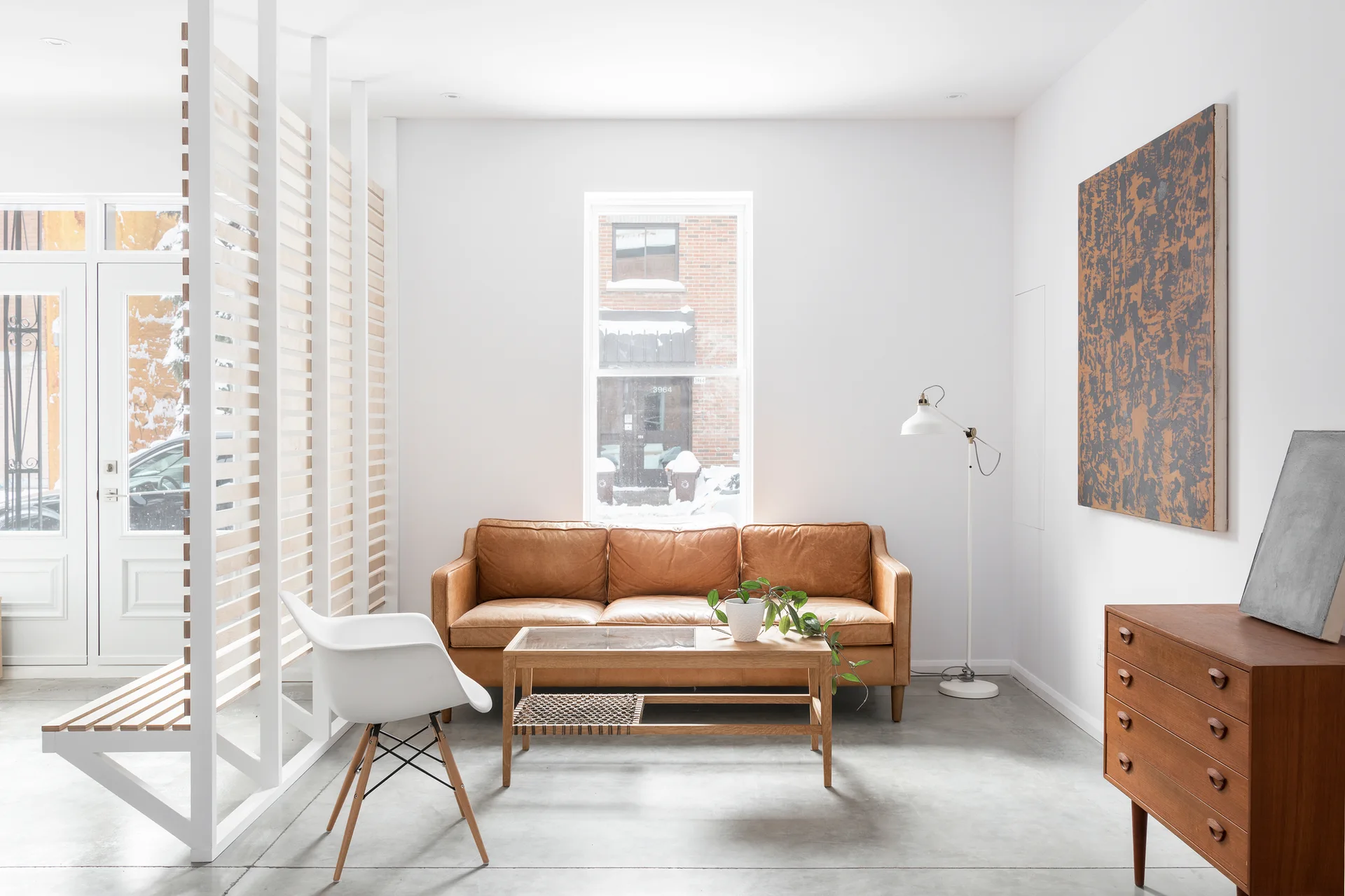 A tan leather sofa sits in a minimalist living room featuring a wooden slat room divider, polished concrete floors, and mid-century wood furniture.