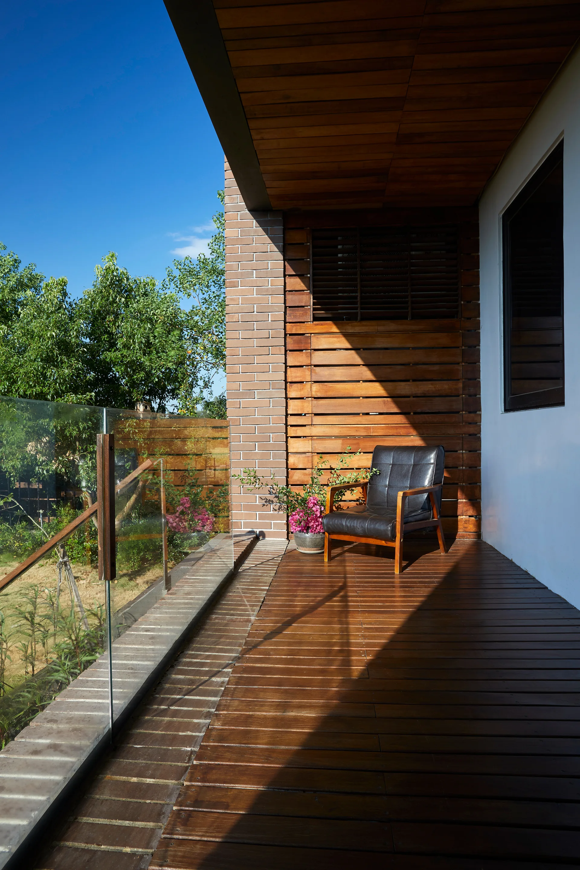 A covered wooden balcony features a black armchair, wood slat walls, a brick column, and a clear glass railing overlooking trees.