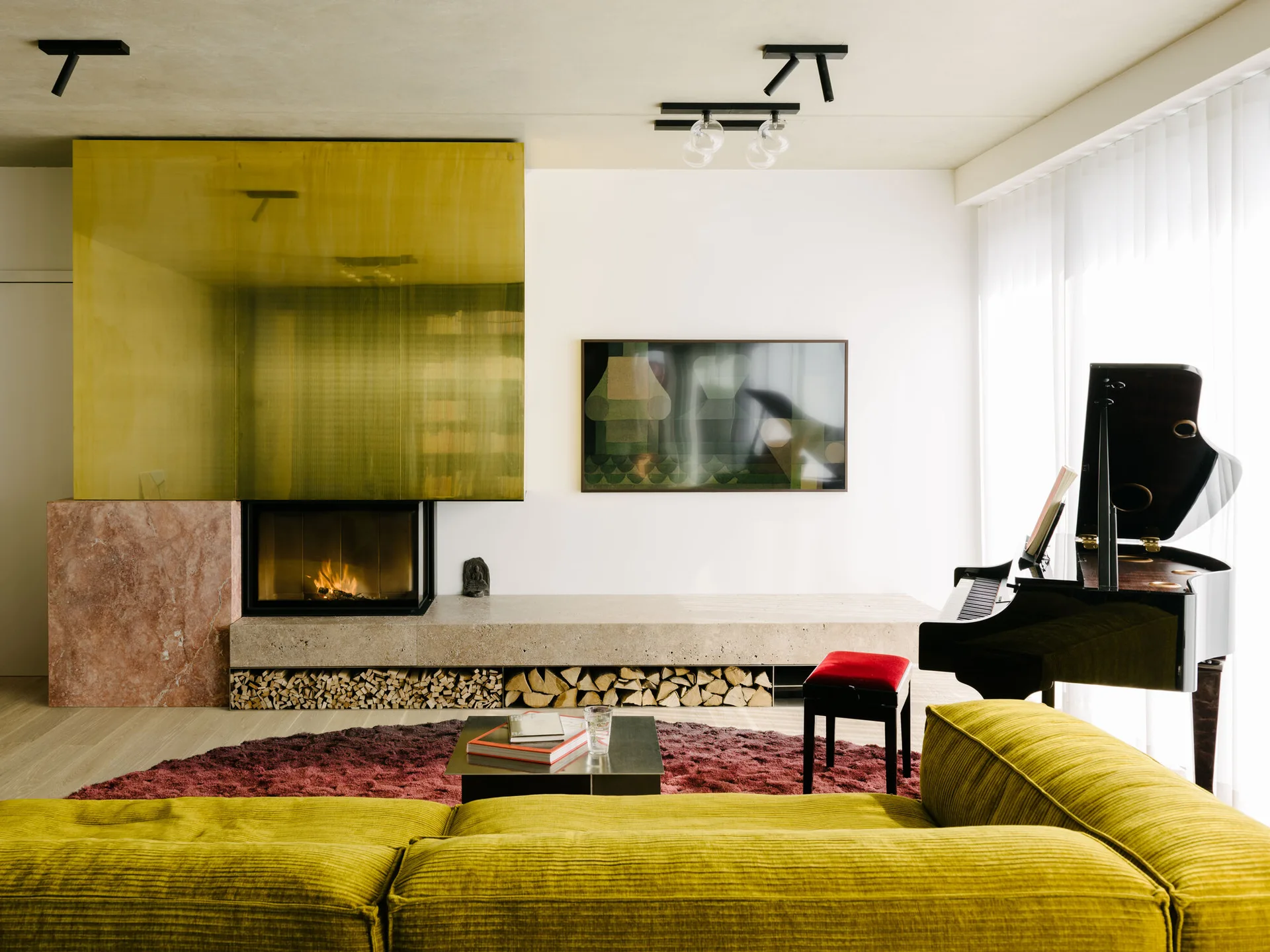 A living room featuring a yellow ribbed sectional, a fireplace with a reflective gold-toned panel, and a black grand piano.