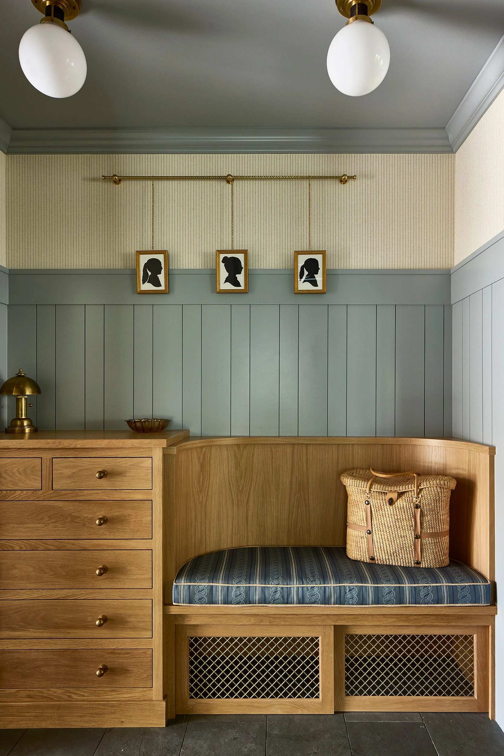 A mudroom corner features a built-in light oak bench and drawers under blue-gray wainscoting with three hanging silhouette portraits.