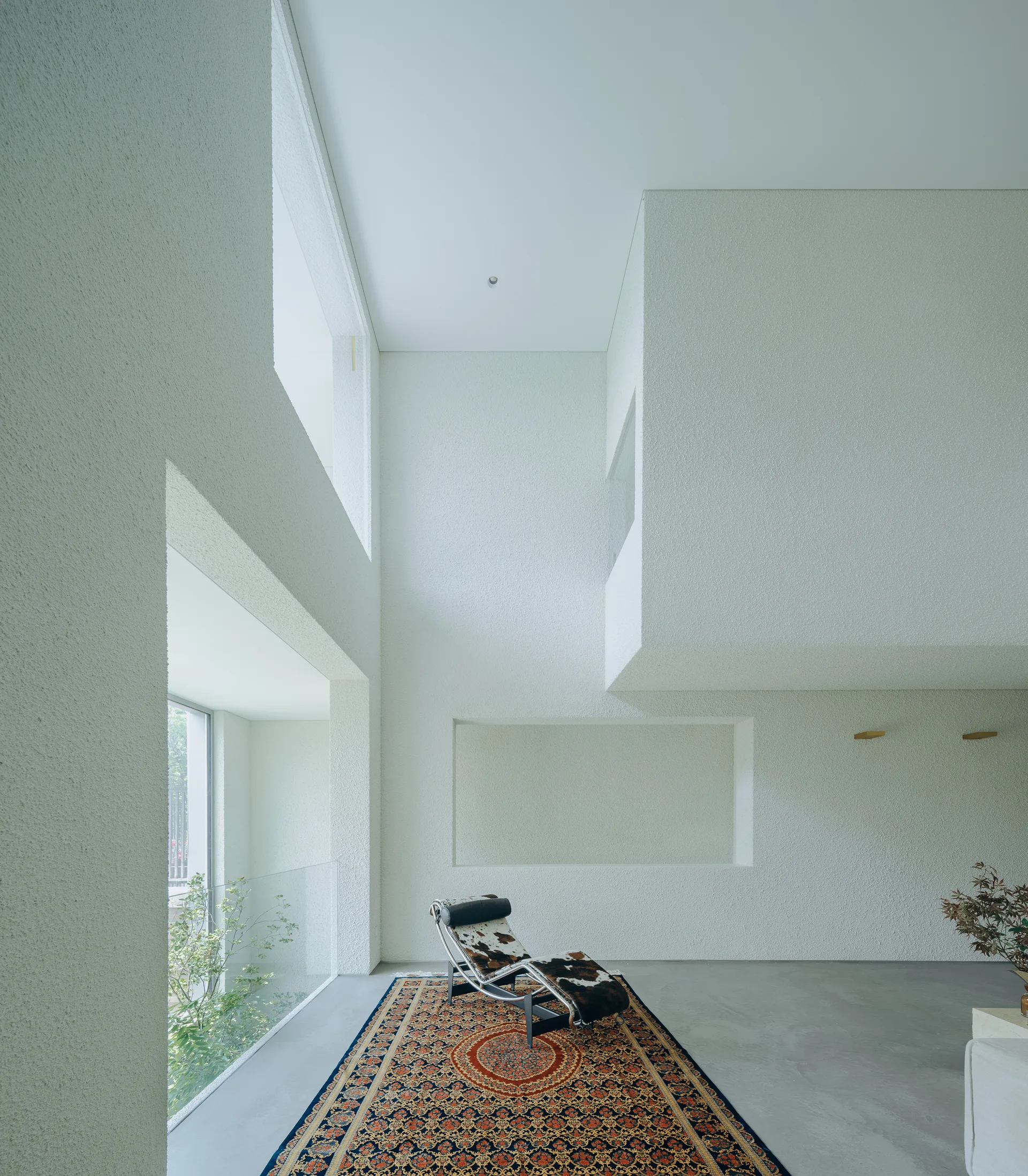 A double-height minimalist room features textured white plaster walls, a gray concrete floor, a patterned oriental rug, and a cowhide chaise lounge.