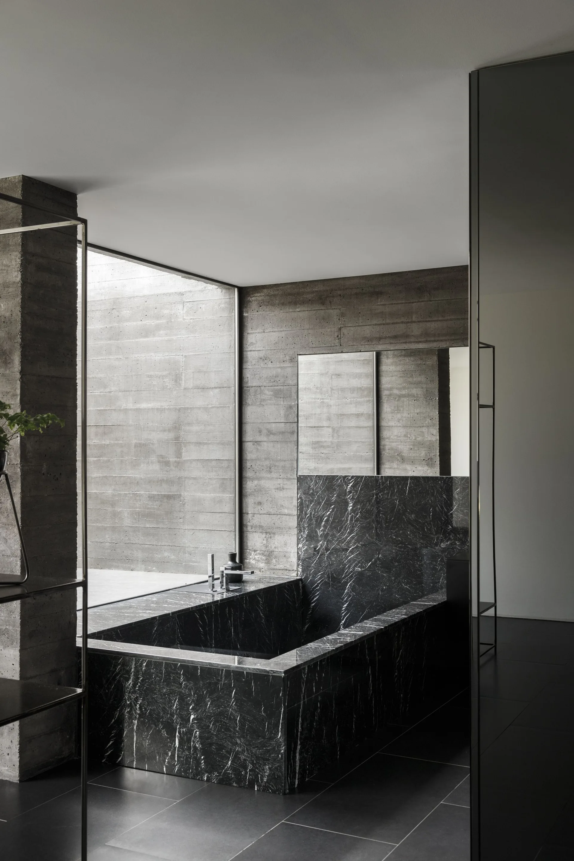 A minimalist bathroom features a black veined marble bathtub set against textured board-formed concrete walls and large windows.