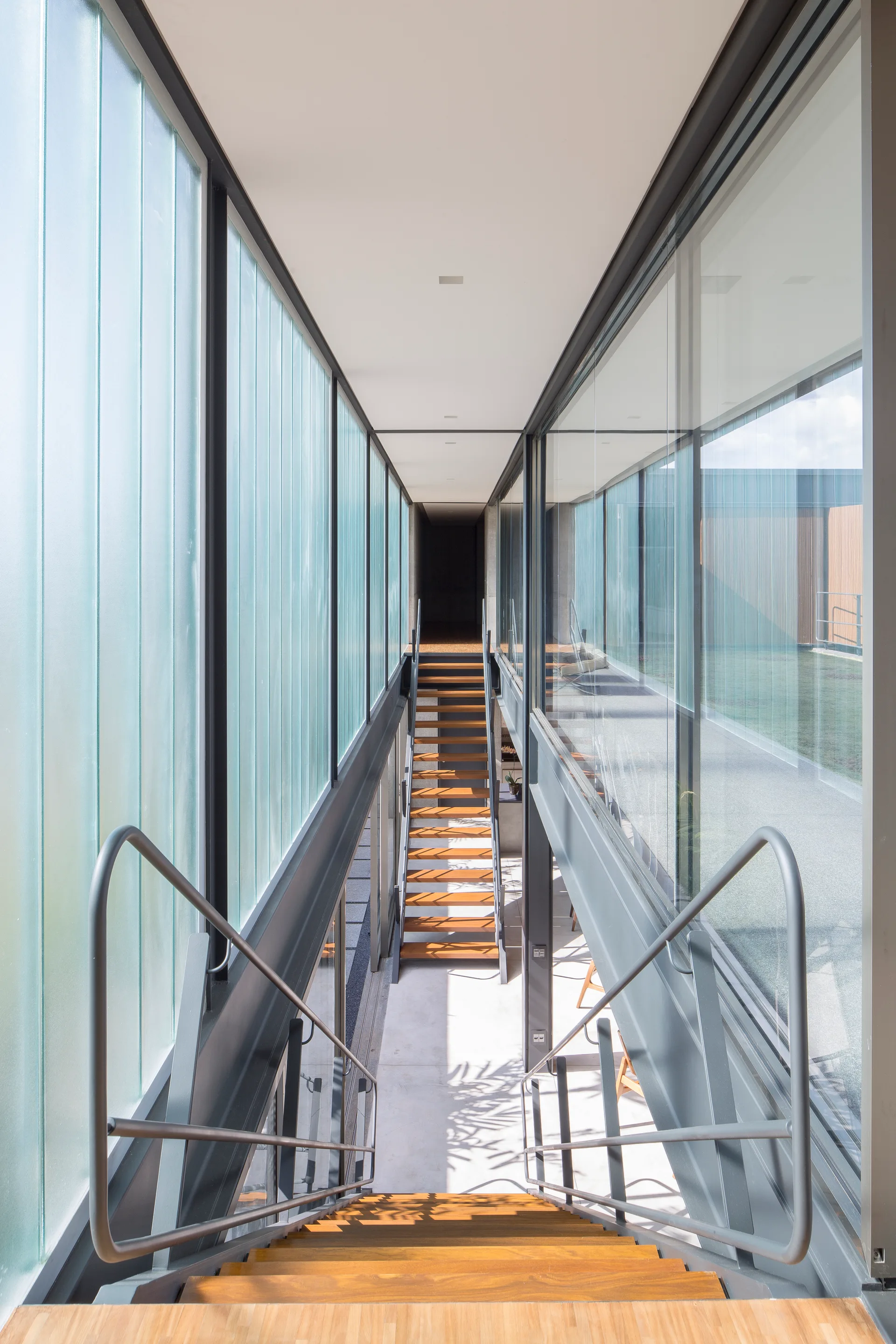 A modern hallway features a central staircase with warm wood treads, metal handrails, and floor-to-ceiling glass partition walls.