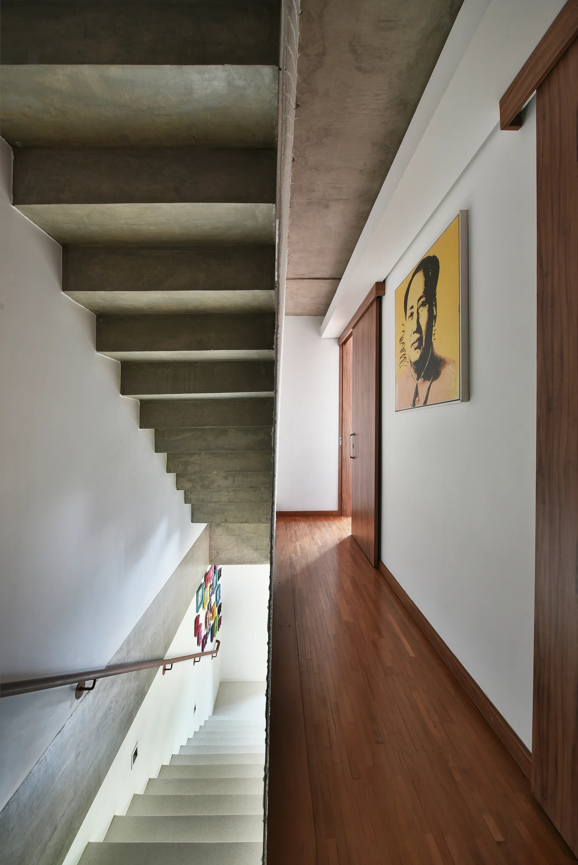A minimalist hallway features warm wood parquet flooring, white walls with sliding wooden doors, and a raw concrete staircase.