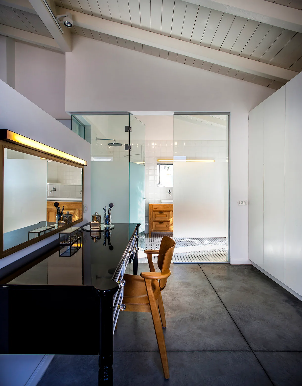 A black vanity table and wooden chair in a dressing room with a concrete floor and a glass partition separating it from a bathroom.
