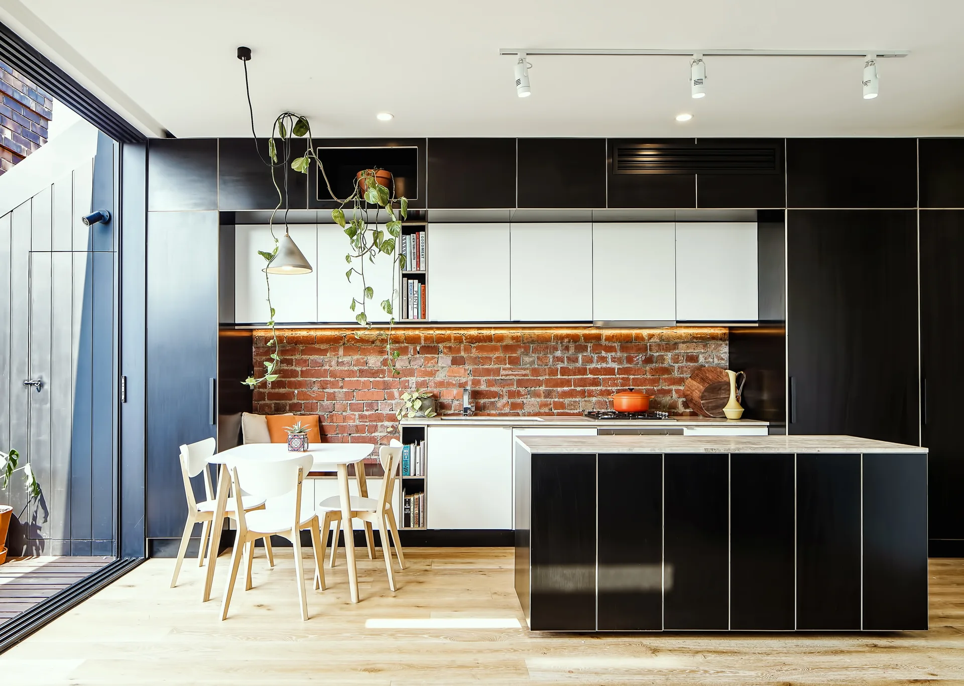 A modern kitchen features black glossy cabinetry, a white dining table with integrated bench seating, and an exposed red brick backsplash.