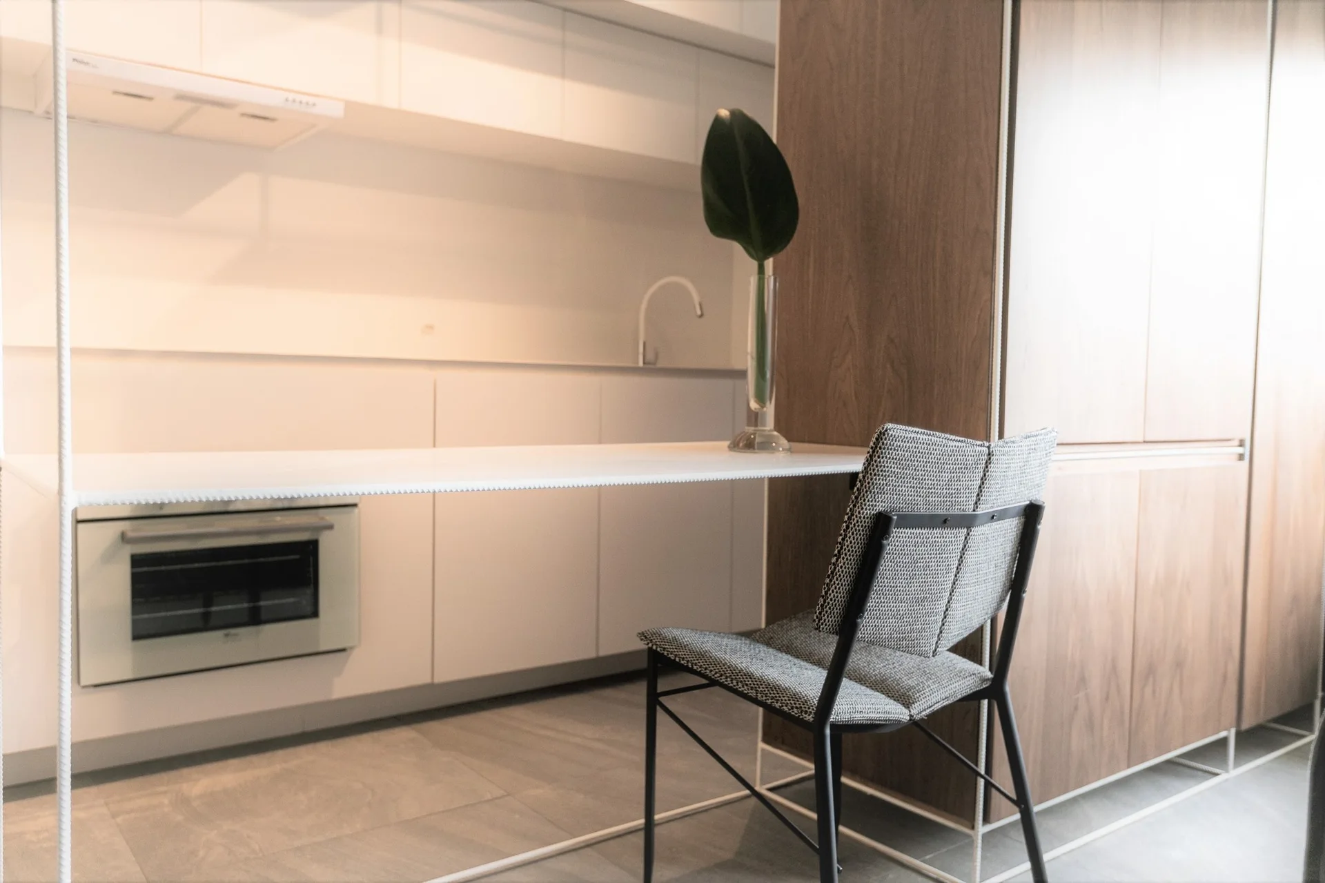 A minimalist kitchen features white flat-front cabinets, a built-in oven, wood-paneled walls, a slim floating counter, and a grey woven fabric chair.