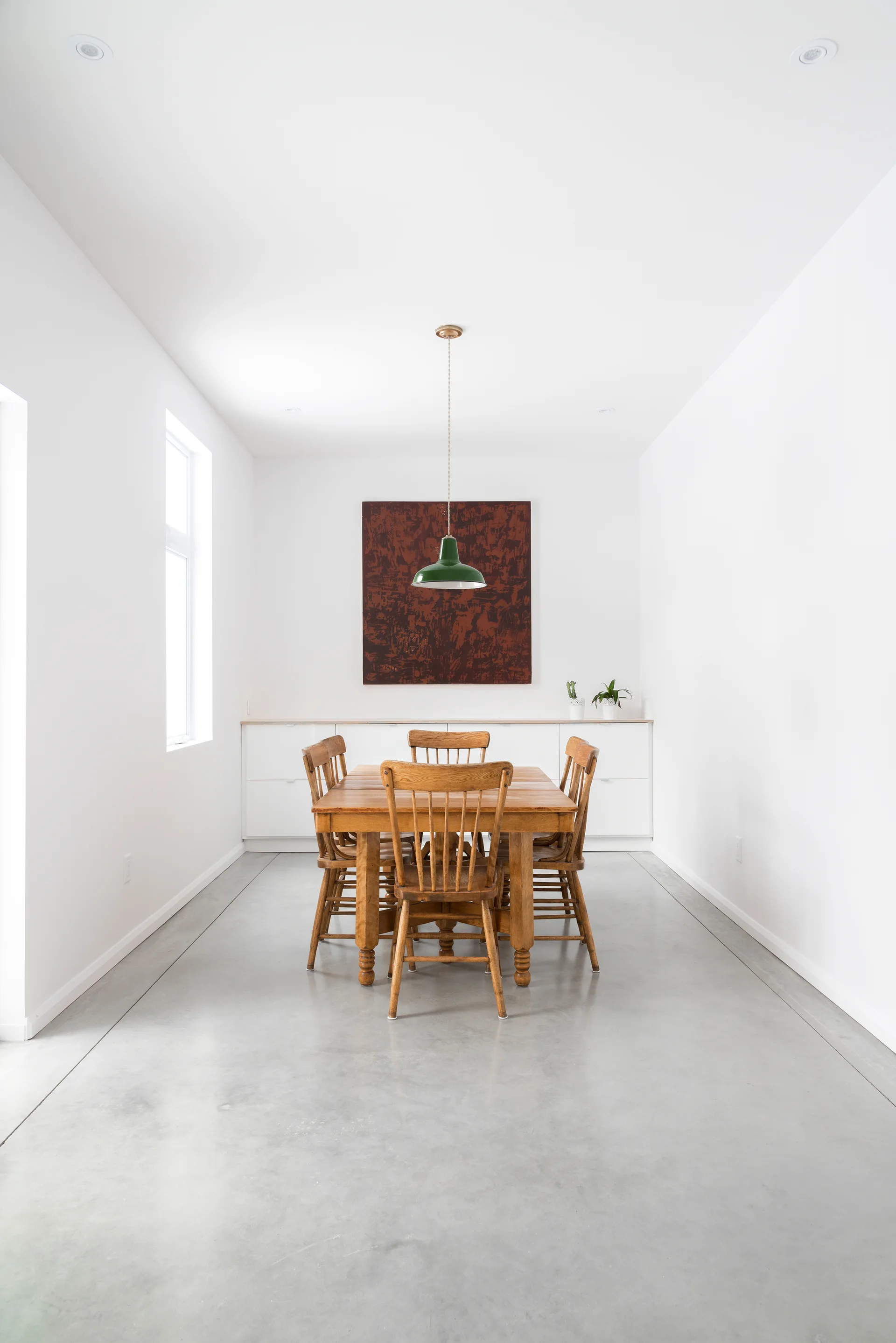 A wooden dining table and chairs sit on a polished concrete floor in a minimalist white room with a green pendant light.