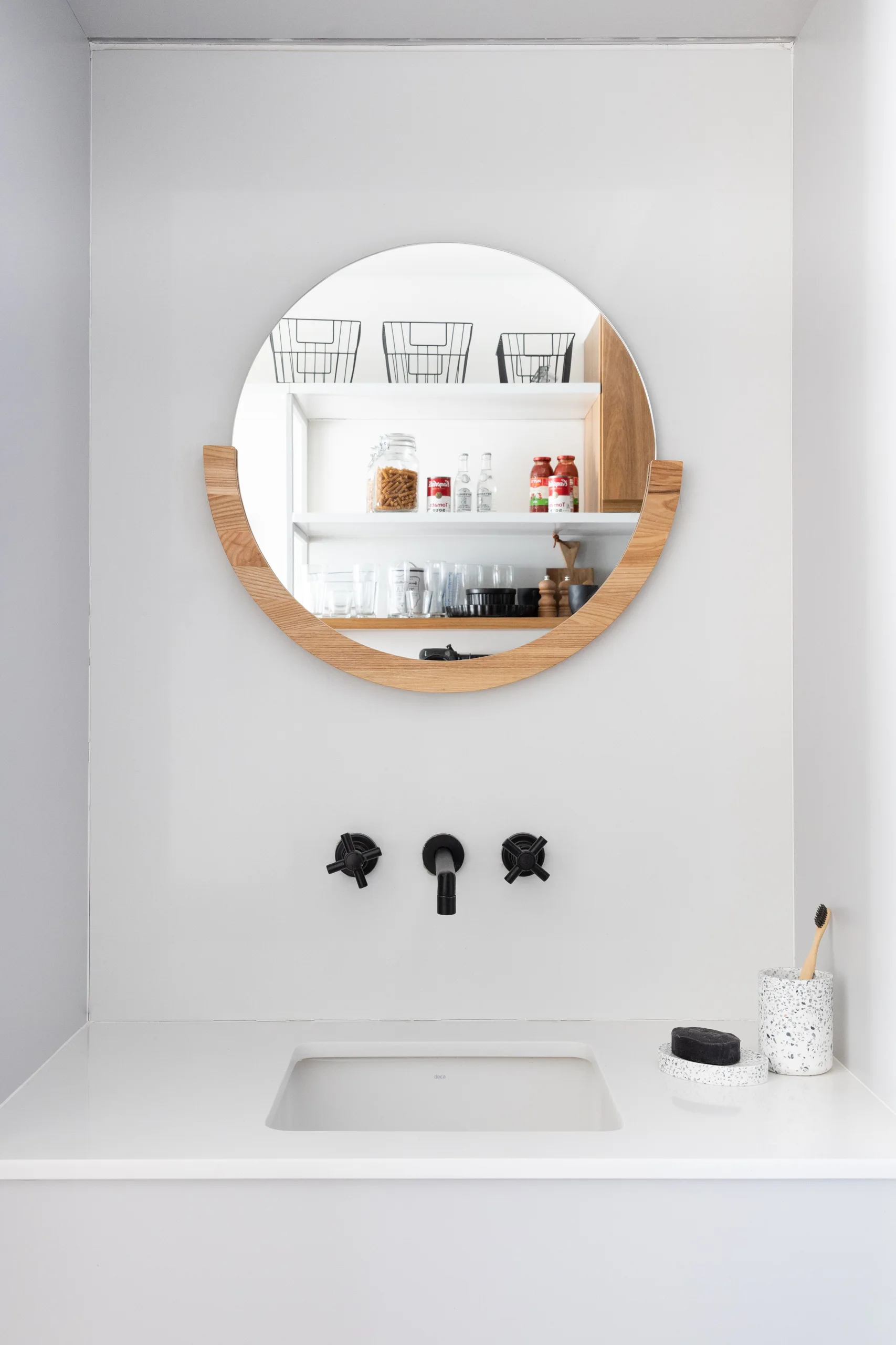 A minimalist white bathroom sink features wall-mounted matte black fixtures below a circular mirror with a curved wooden lower frame.