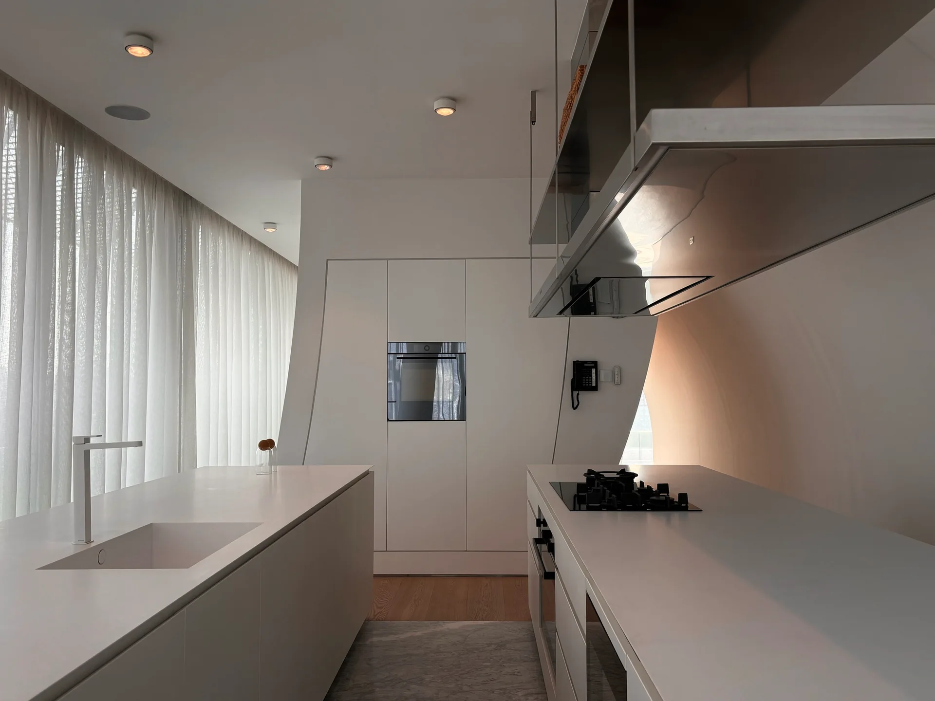 A minimalist white kitchen features a central island with a built-in sink, a metal range hood, and a unique curved architectural wall.