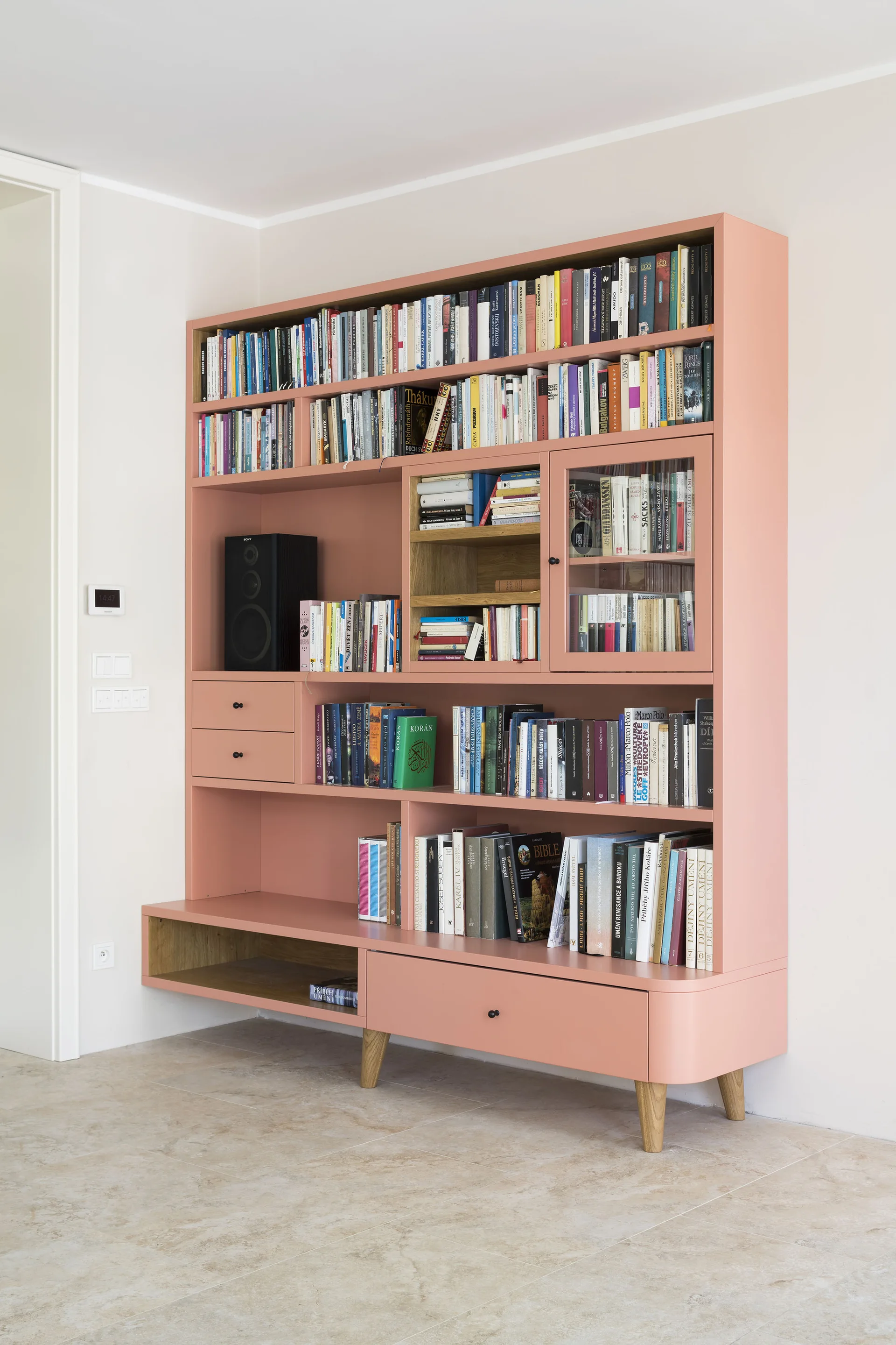 A large matte pink bookshelf filled with books stands against a white wall on a light stone tile floor.