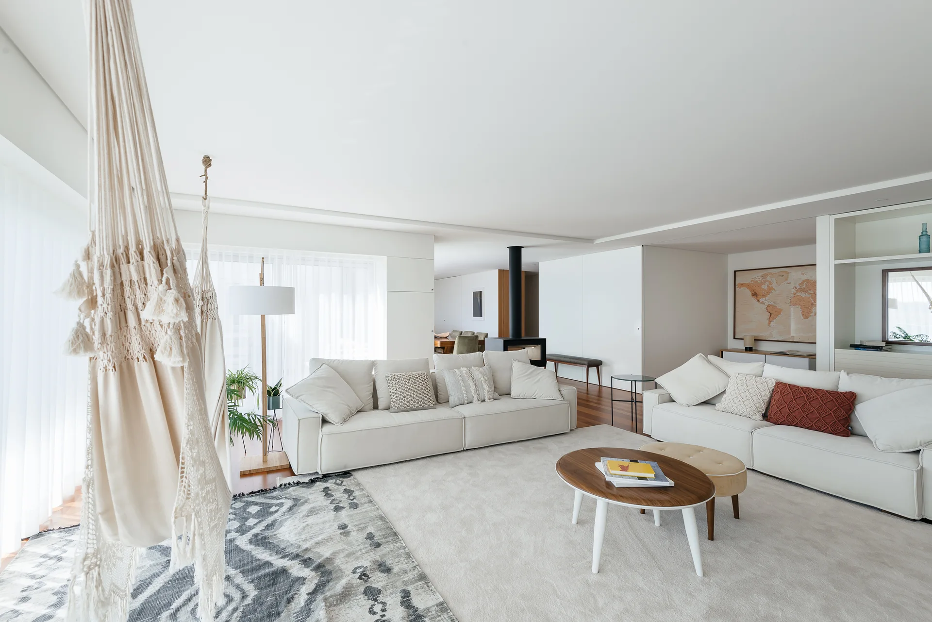 A minimalist white living room features two cream sofas, a round wooden coffee table, a black fireplace, and a macrame hanging chair in the foreground.