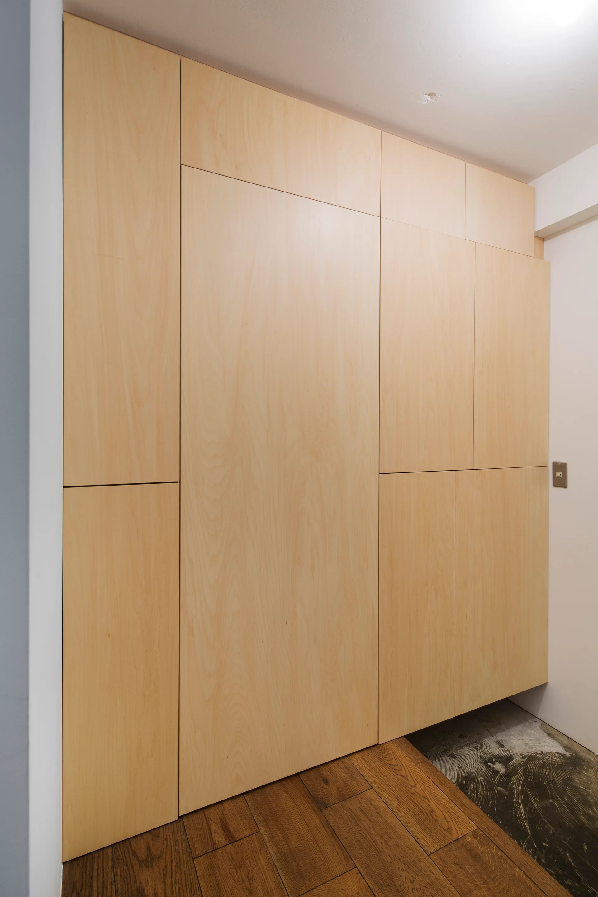 A full-height custom built-in closet with flat-panel blonde wood doors and a handleless design stands next to a white wall and wood plank flooring.