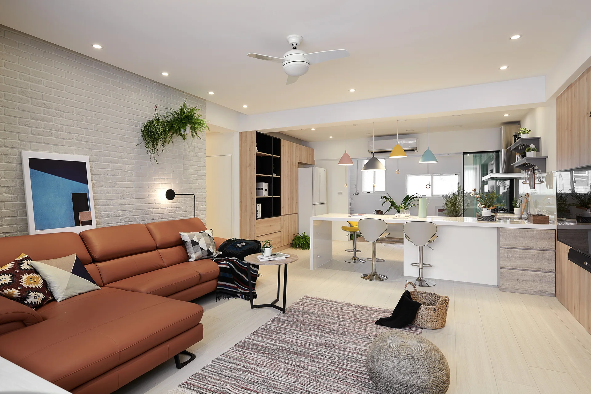 An open-plan living room and kitchen features a brown leather sectional sofa, a white-painted brick wall, and a kitchen island with colorful pendant lights.