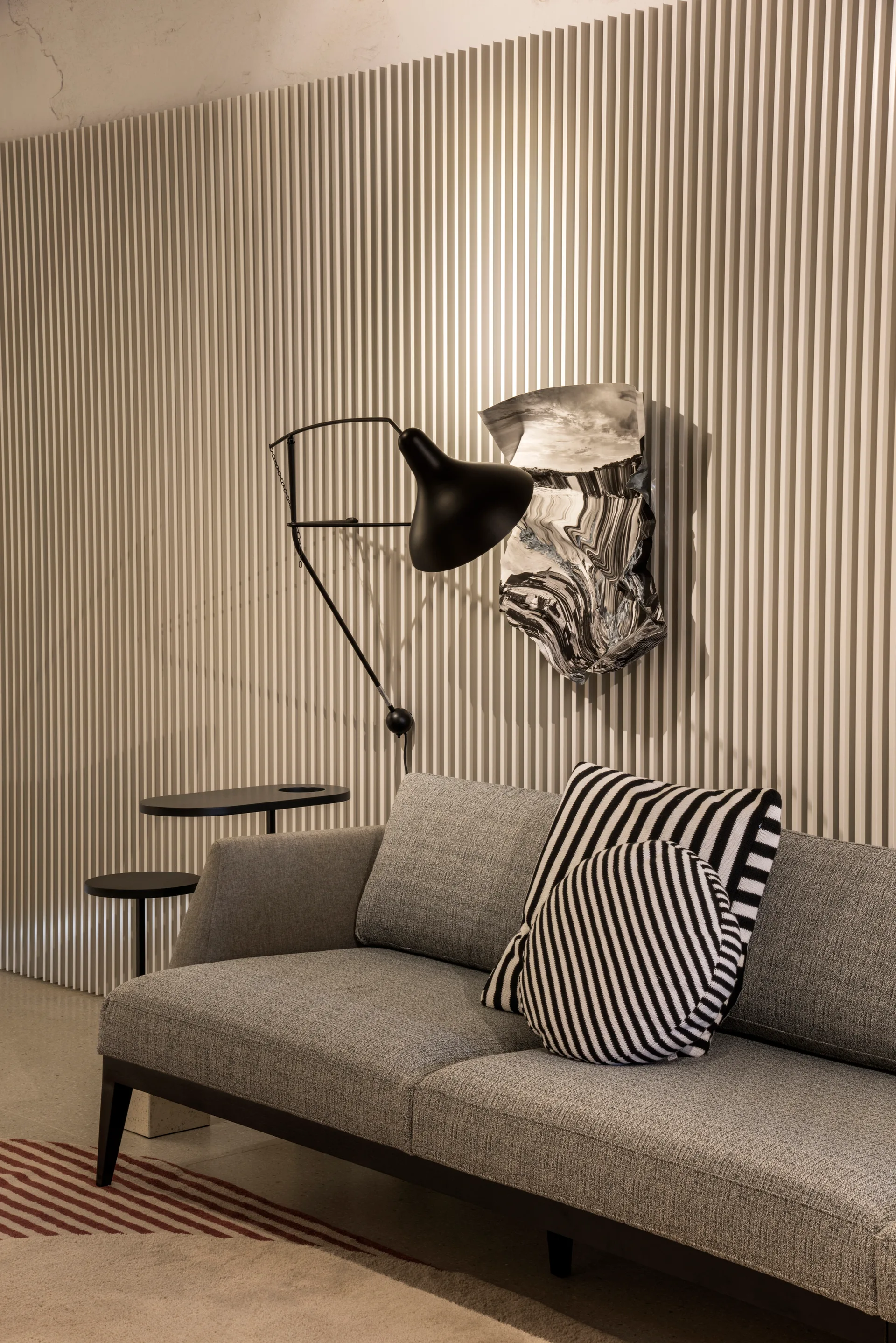 A gray upholstered sofa with striped black and white pillows sits against a beige vertical slatted wall near a black swing-arm lamp.