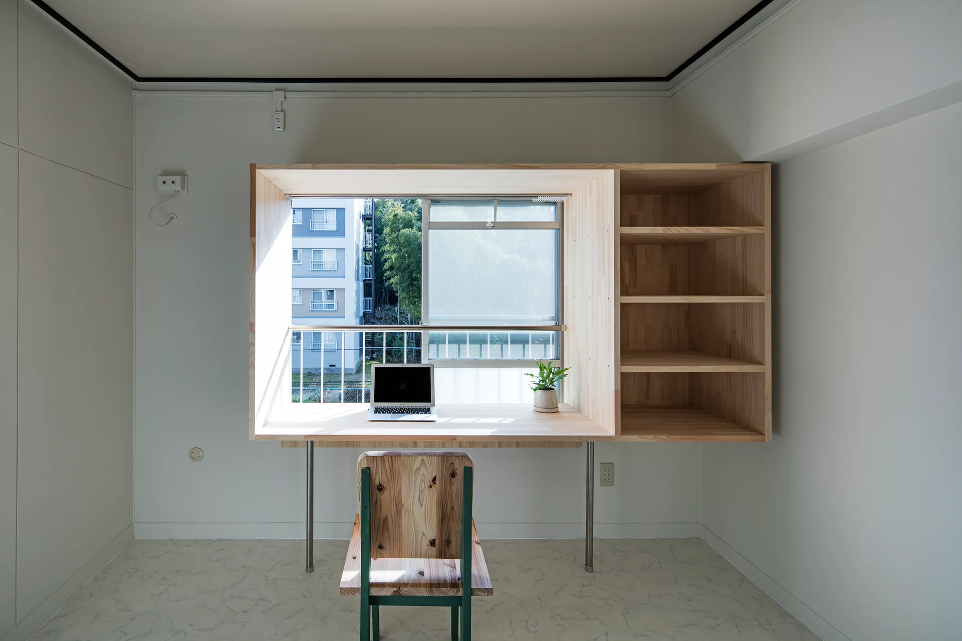 A minimalist home office features a natural light wood desk and shelving unit built around a window with a matching wooden chair in front.