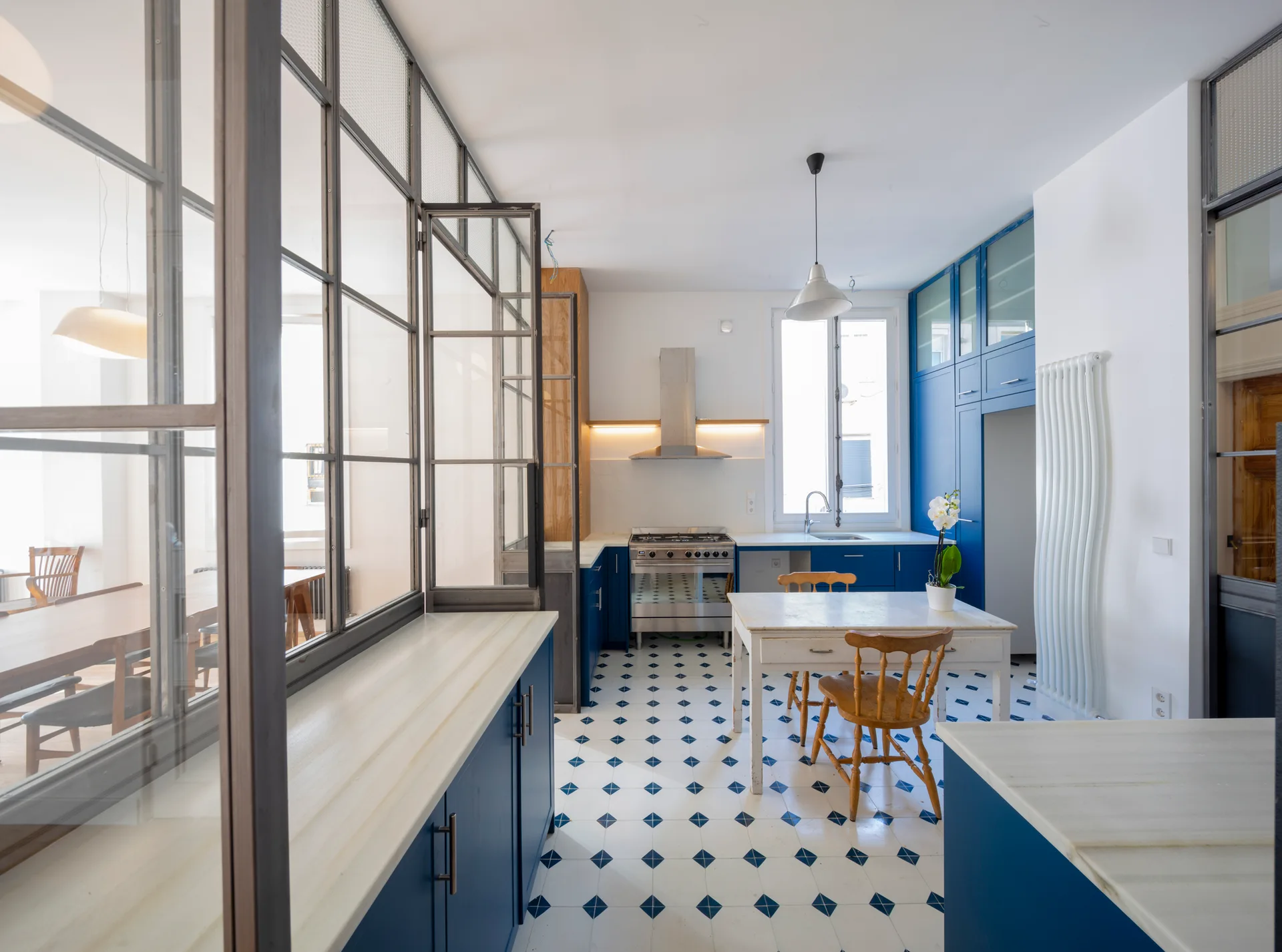 A bright kitchen features blue cabinetry, a diamond-patterned tile floor, a central wooden dining table, and a steel-framed glass partition wall.