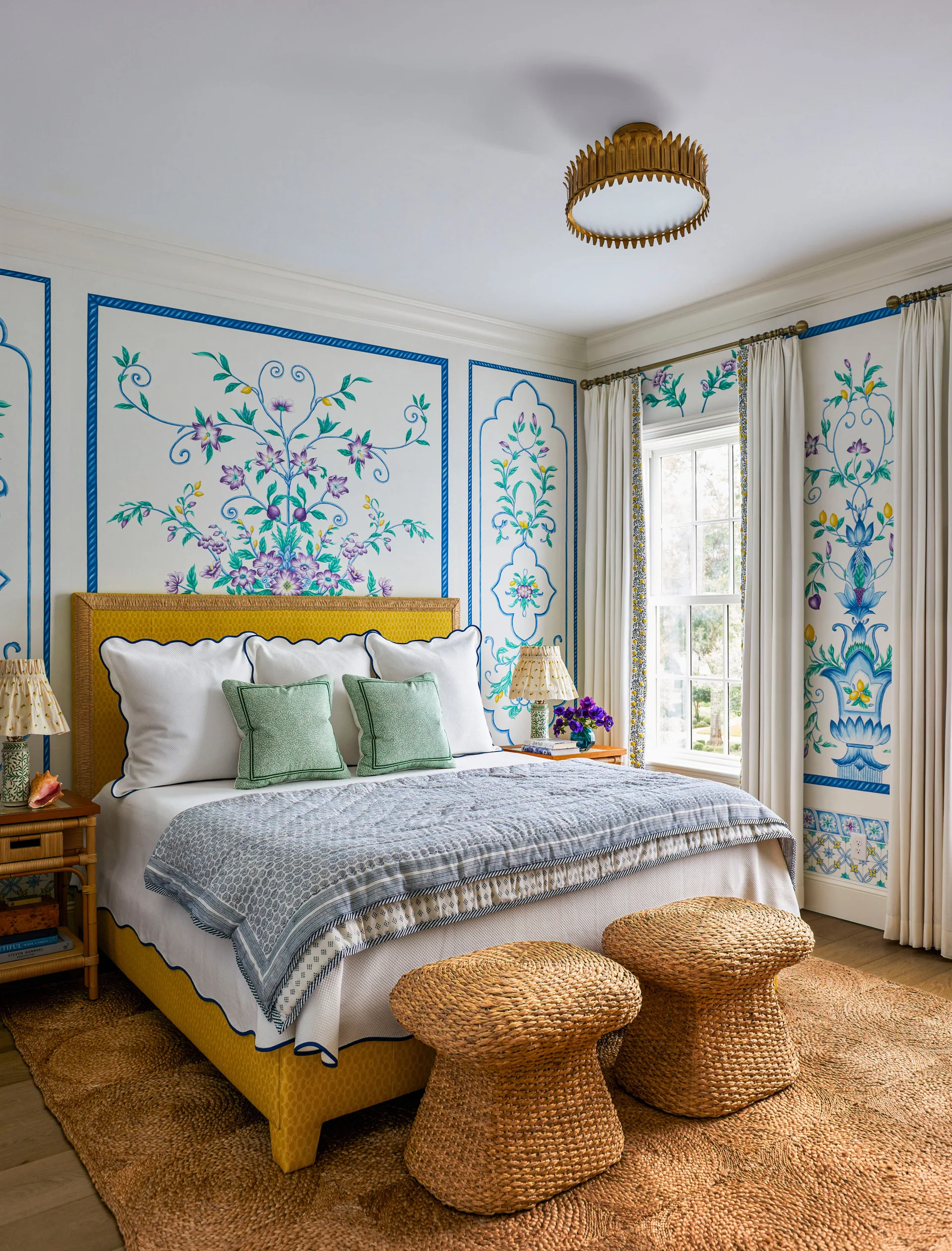 A traditional bedroom featuring a yellow upholstered bed, colorful floral wall murals framed by blue trim, and two woven wicker stools.