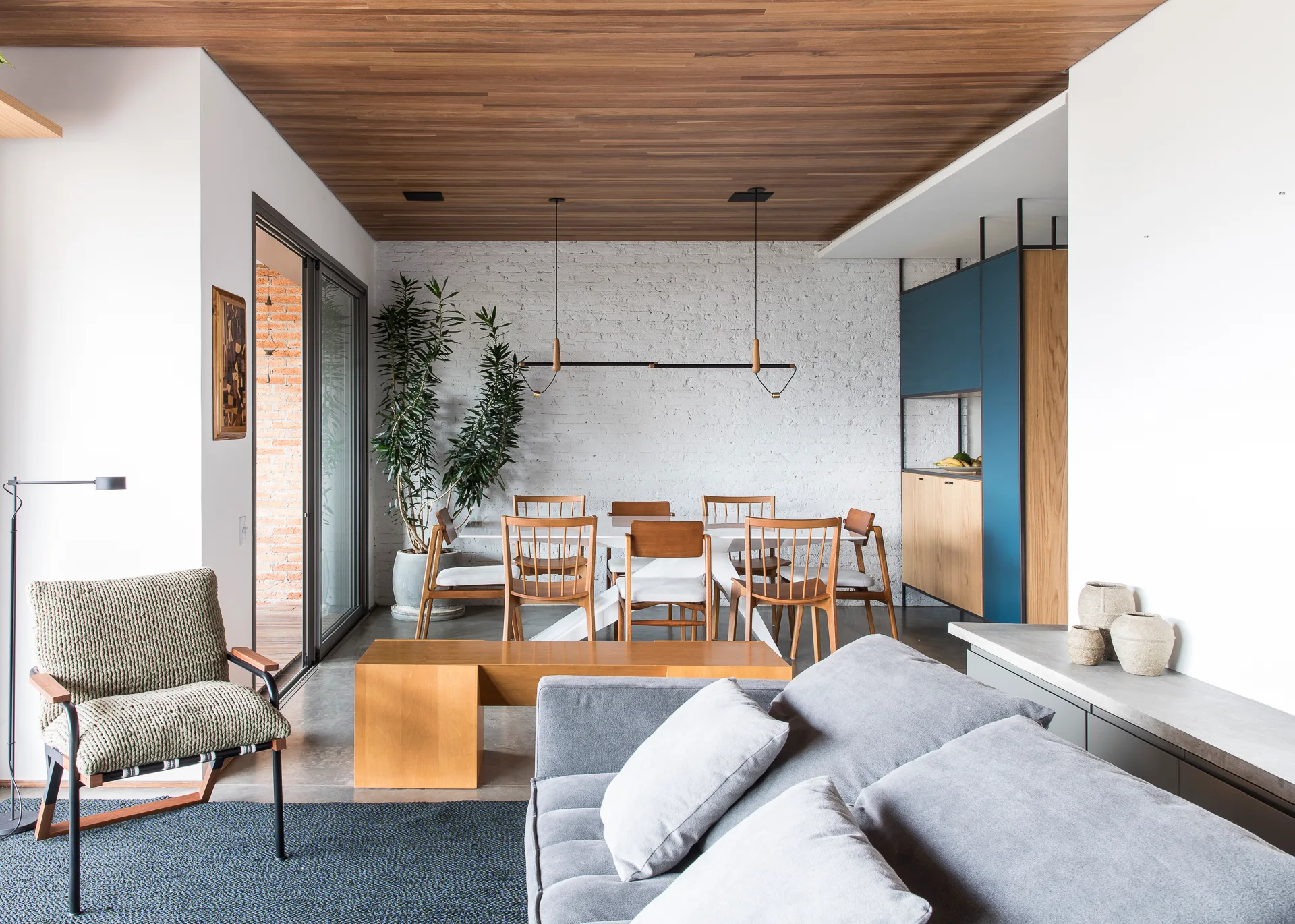 A contemporary open-plan dining and living space featuring a grey sofa, a wooden dining table, a white brick wall, and a wood plank ceiling.