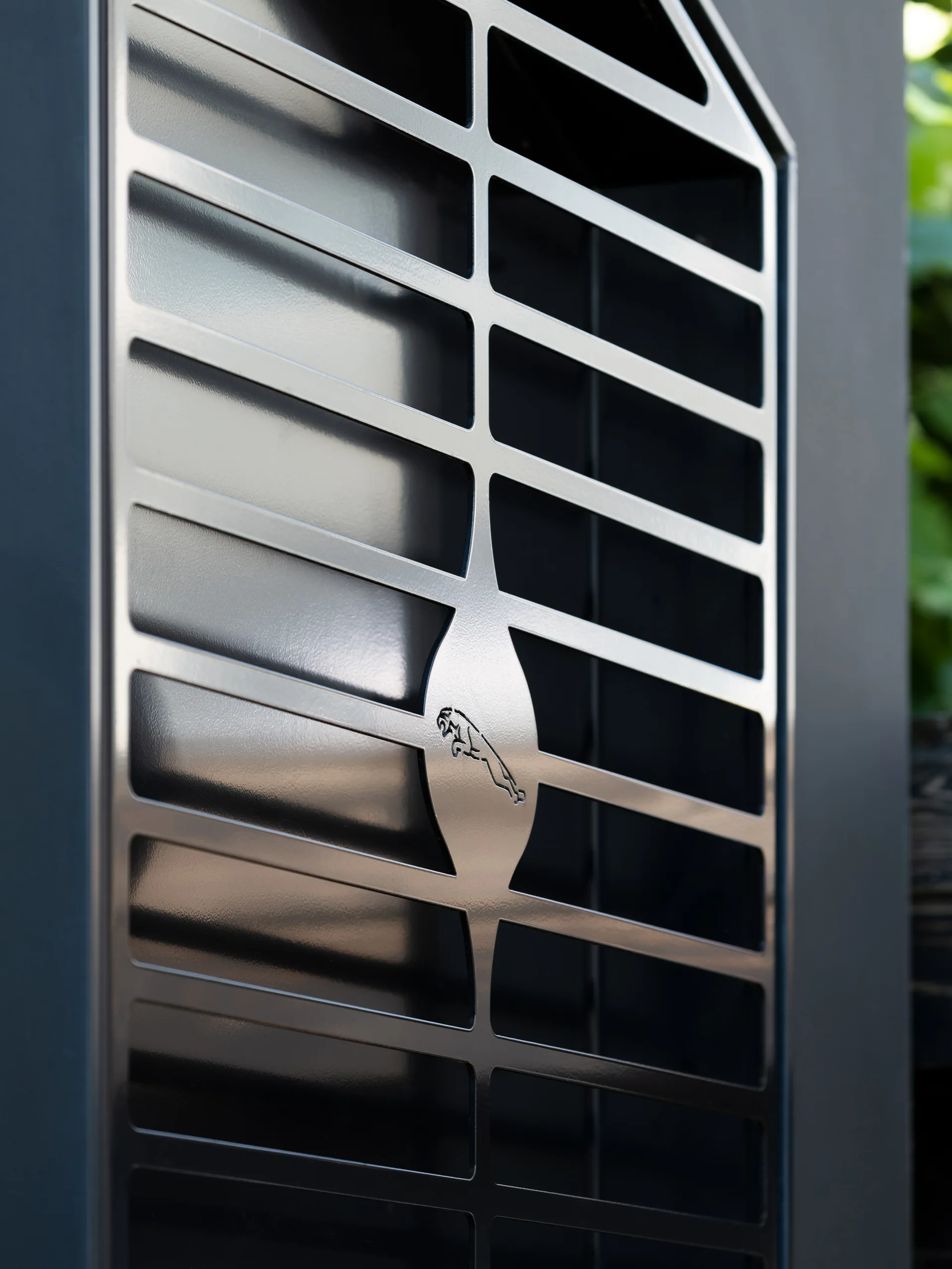 A close-up of a dark metallic architectural grille featuring a geometric slotted grid pattern and a central leaping cat emblem.
