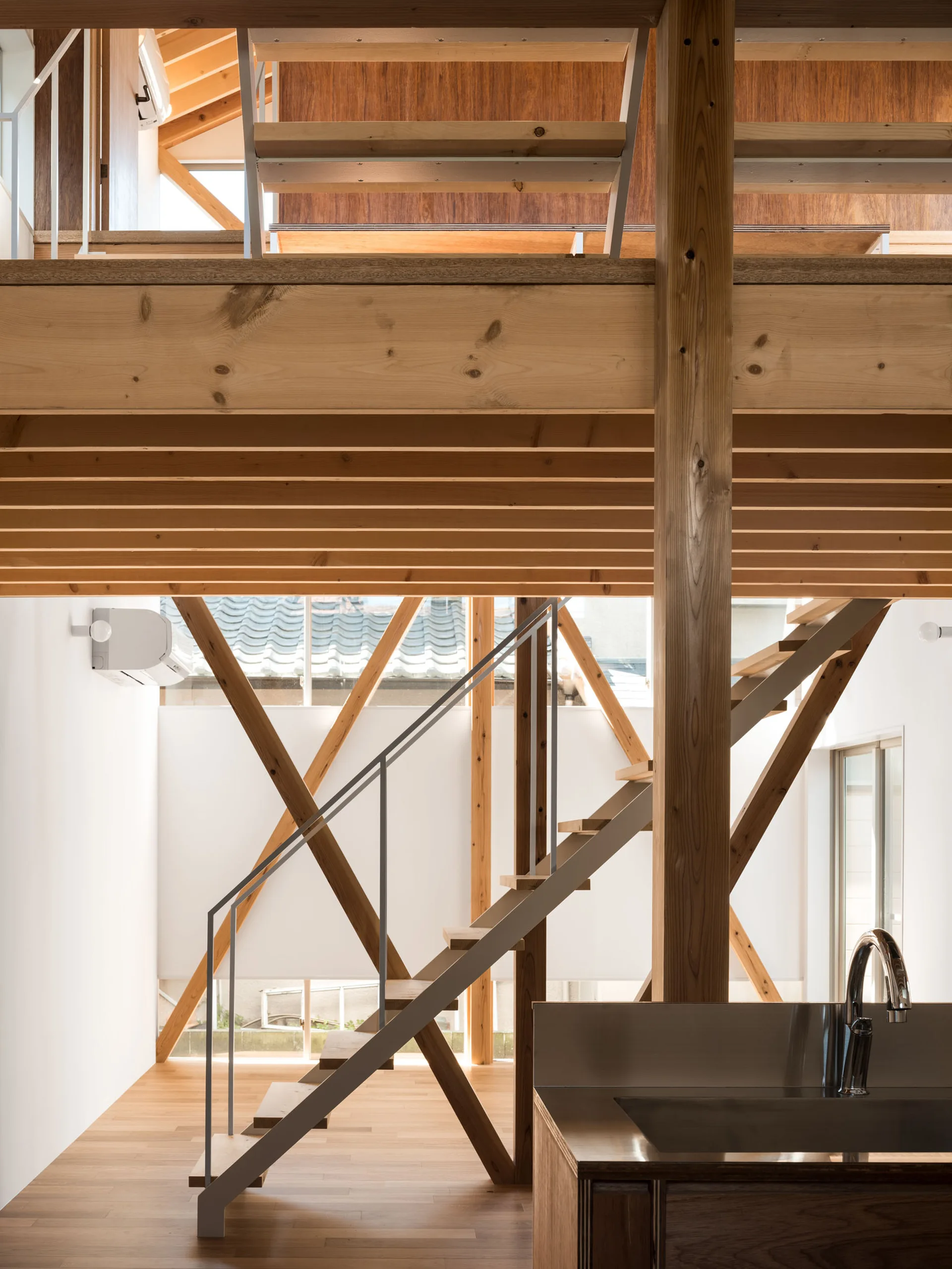 A bright interior featuring a minimalist floating staircase framed by exposed light wood timber posts and beams, with a kitchen sink in the foreground.