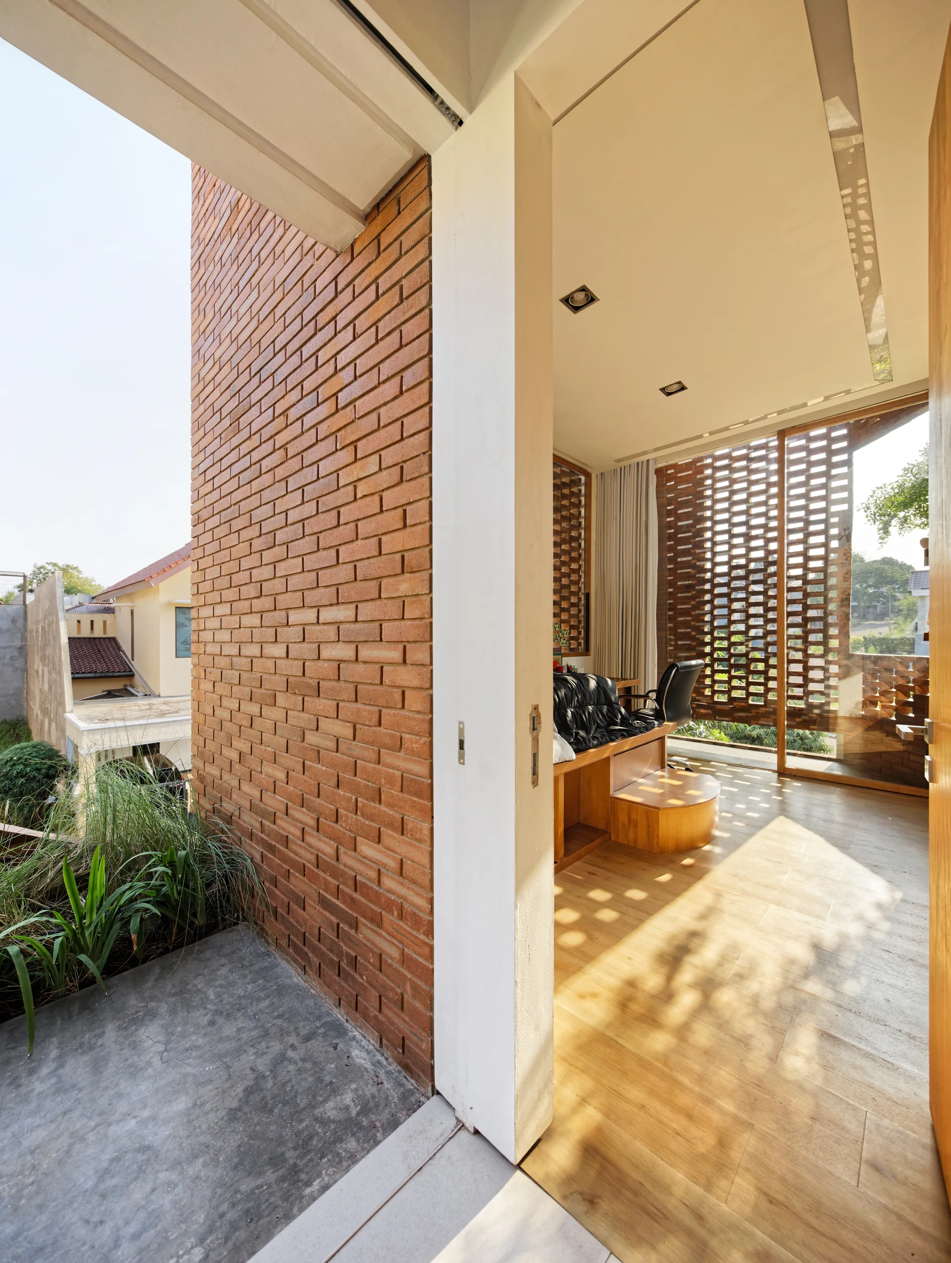 A doorway reveals a sunlit home office with wood floors, a wooden desk, and a perforated brick screen that casts geometric shadows.