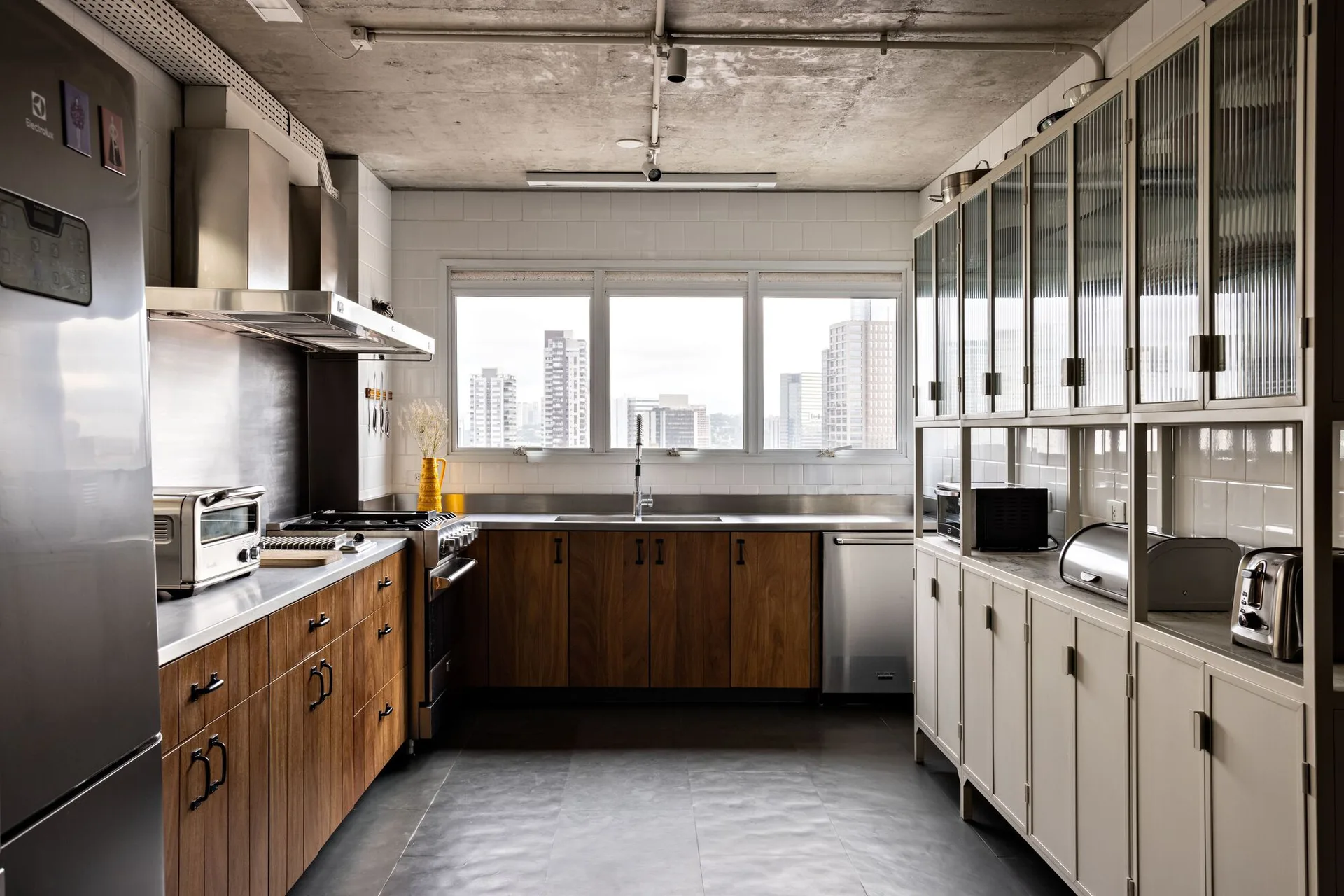 A galley kitchen features wood base cabinets, a light beige hutch with fluted glass doors, and an exposed raw concrete ceiling with exposed pipes.
