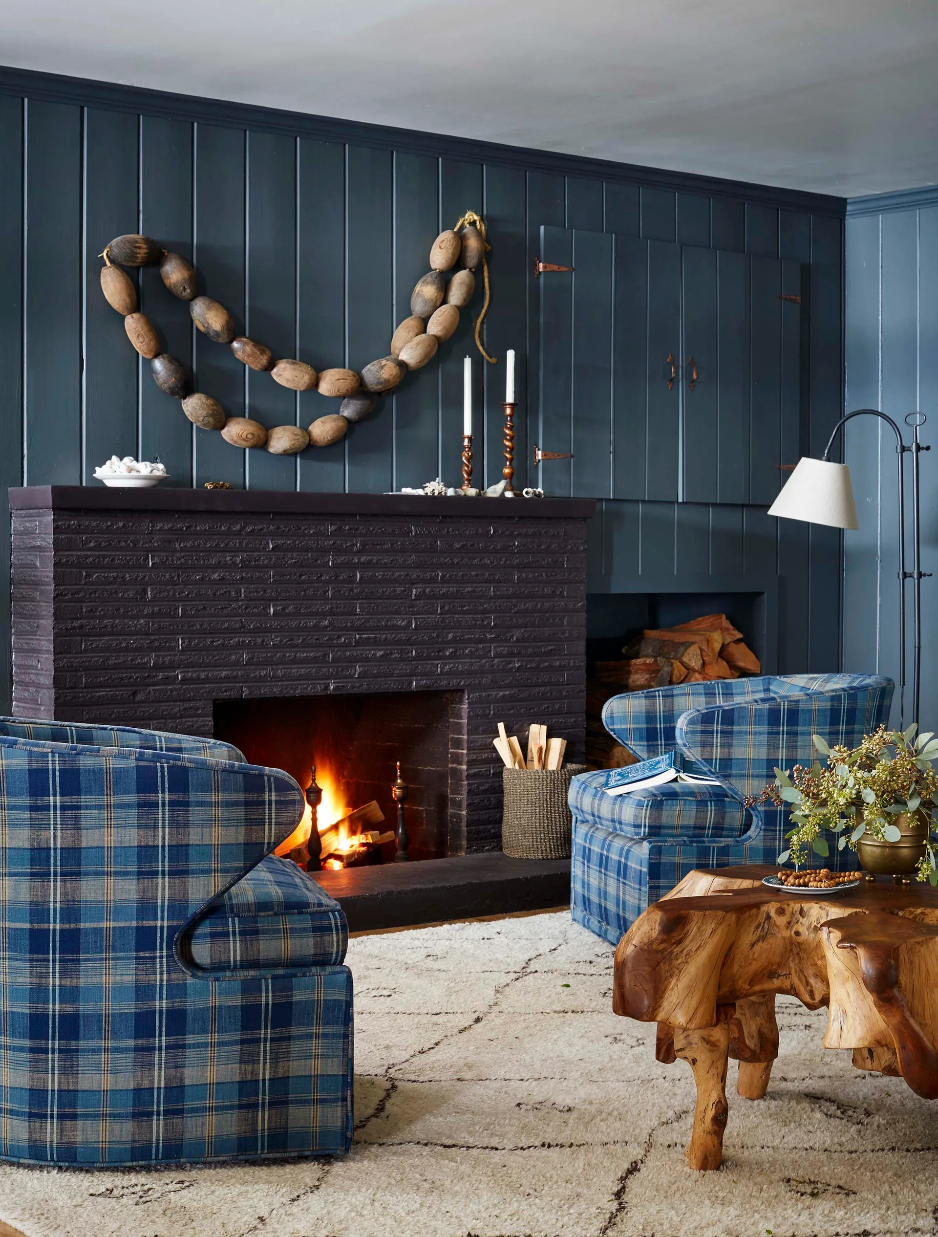 Two blue plaid armchairs sit before a burning brick fireplace set against navy vertical wall paneling, with a live-edge wood coffee table in front.