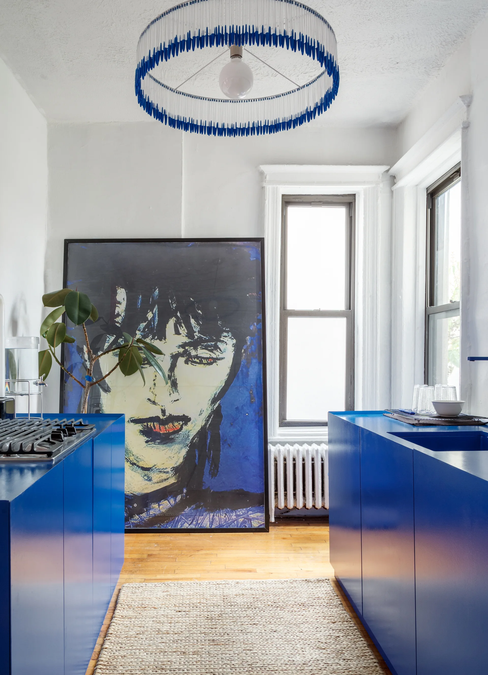 A kitchen features bright blue cabinets, a woven runner rug, and a large framed portrait leaning on the floor between two windows.