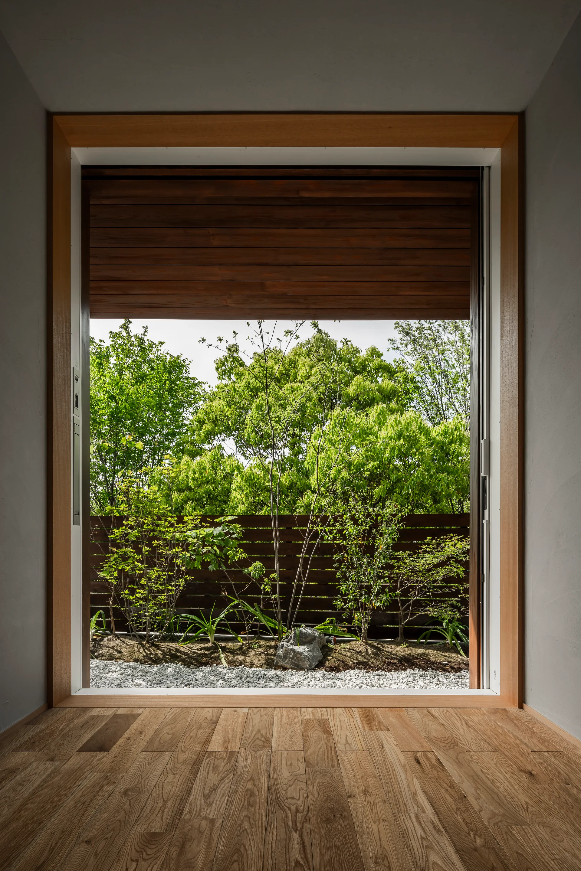 A minimalist room with light wood floors features a large natural wood frame looking out onto a garden with white gravel and green trees.