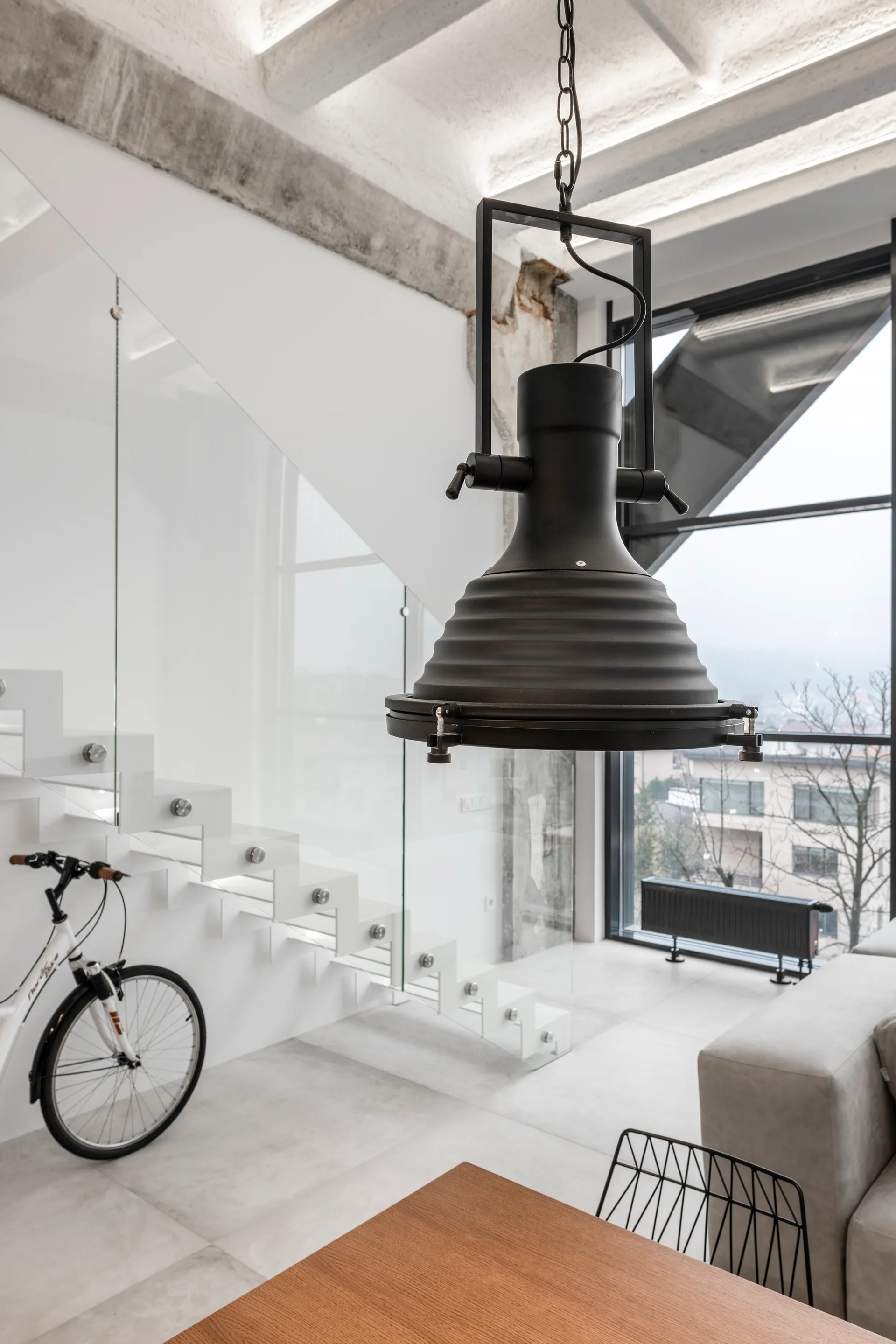 A modern loft interior features a white floating staircase with a glass railing, a large black industrial pendant light, and a concrete ceiling.