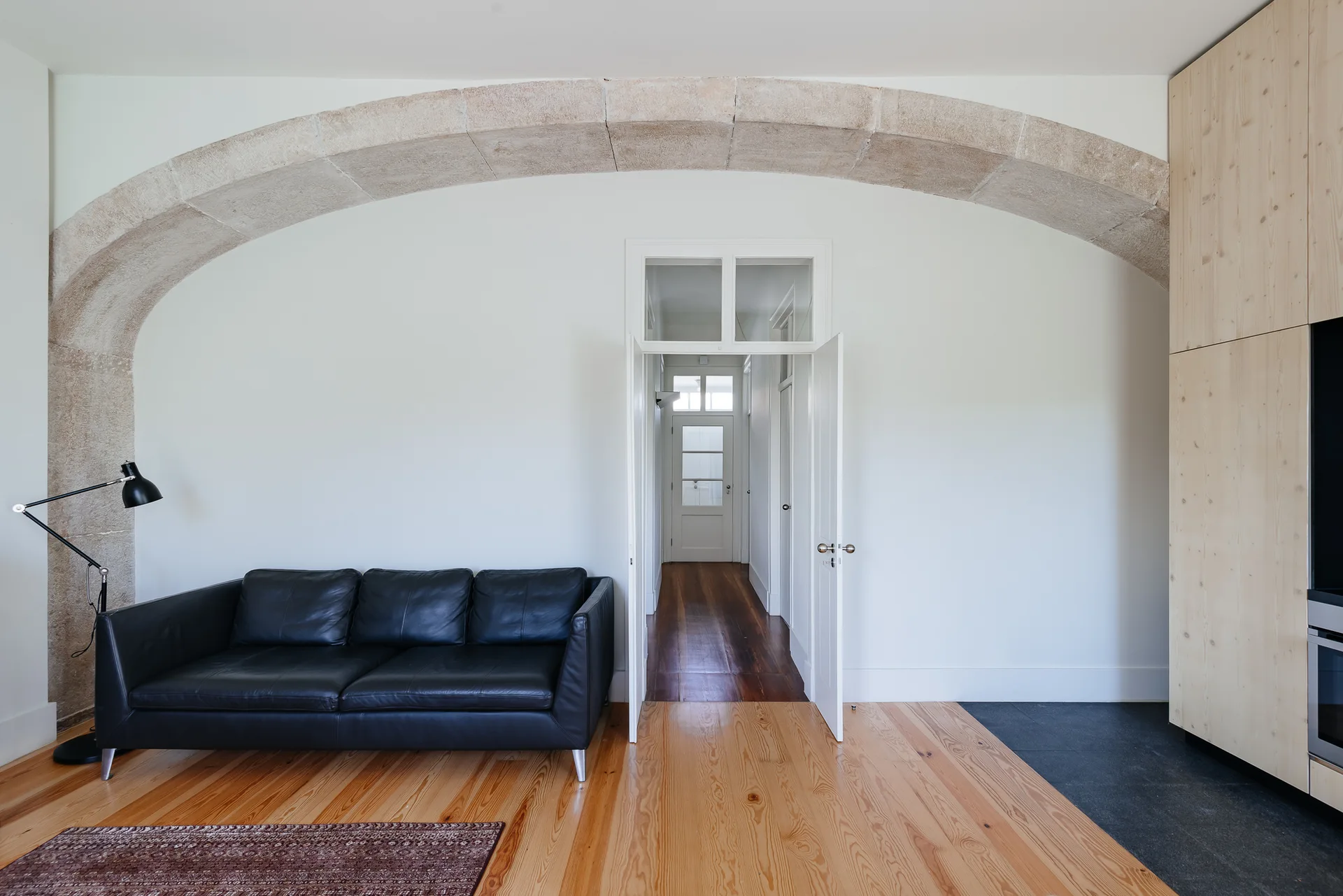 A minimalist room with a black leather sofa beneath a large stone archway and light wood flooring.