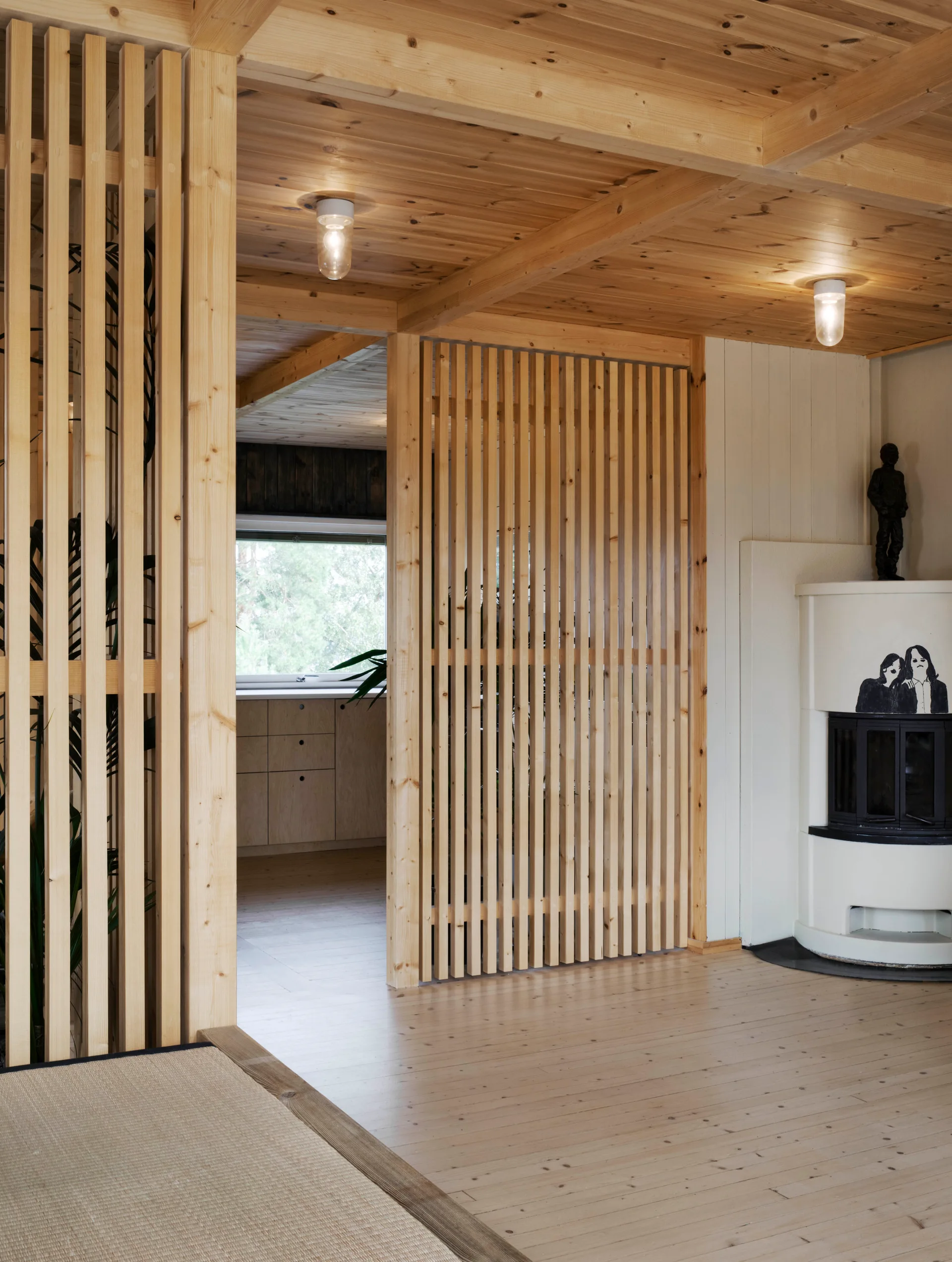 An interior space features a vertical light wood slat partition separating the living room from a kitchen with wood cabinets and a white fireplace.